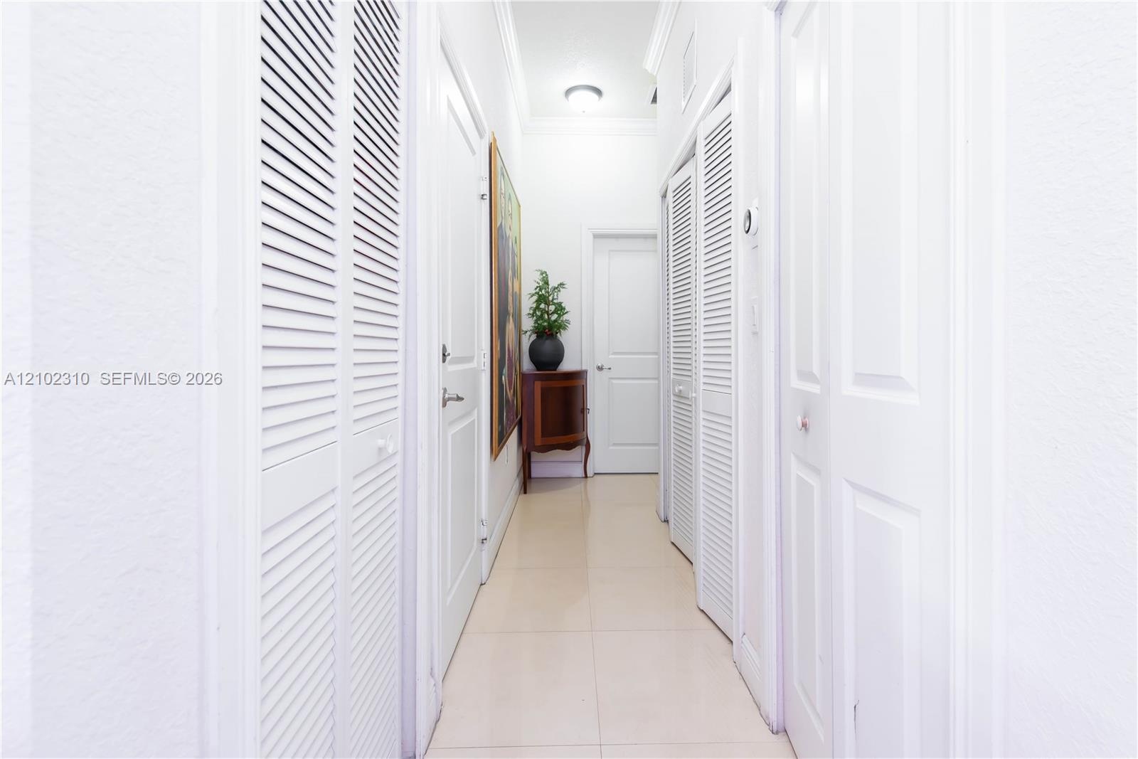 Hallway featuring neutral large-format tile flooring, multiple sets of white louvered-style closet doors, solid-panel interior doors, and crown moldin