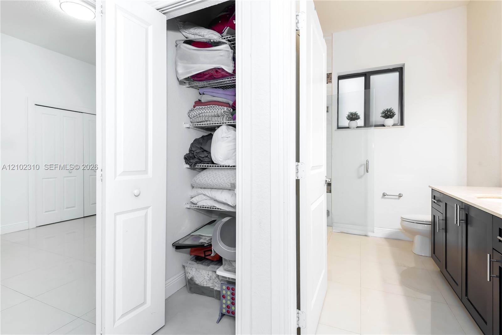 Dark wood-finish cabinetry, light-toned countertop, wire shelving unit, white tile flooring, and a glass-enclosed shower