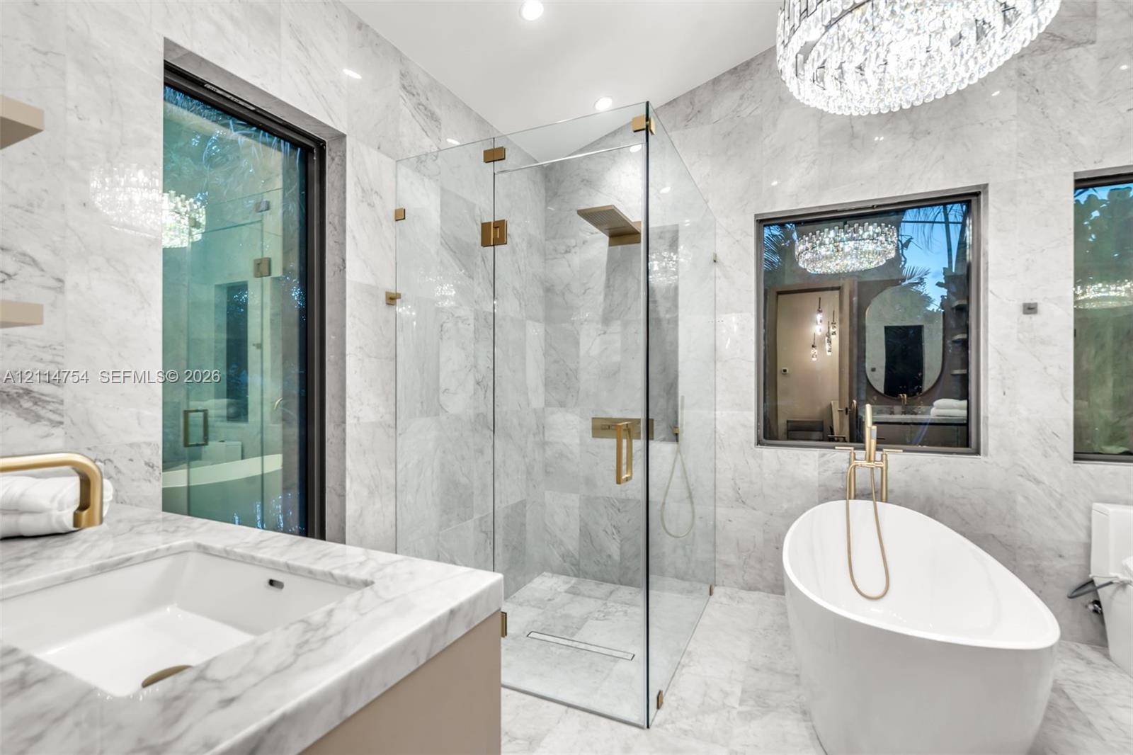 Stone tile wall and floor surfaces, glass-enclosed walk-in shower with gold-tone fixtures, freestanding soaking tub with floor-mounted faucet, and cus