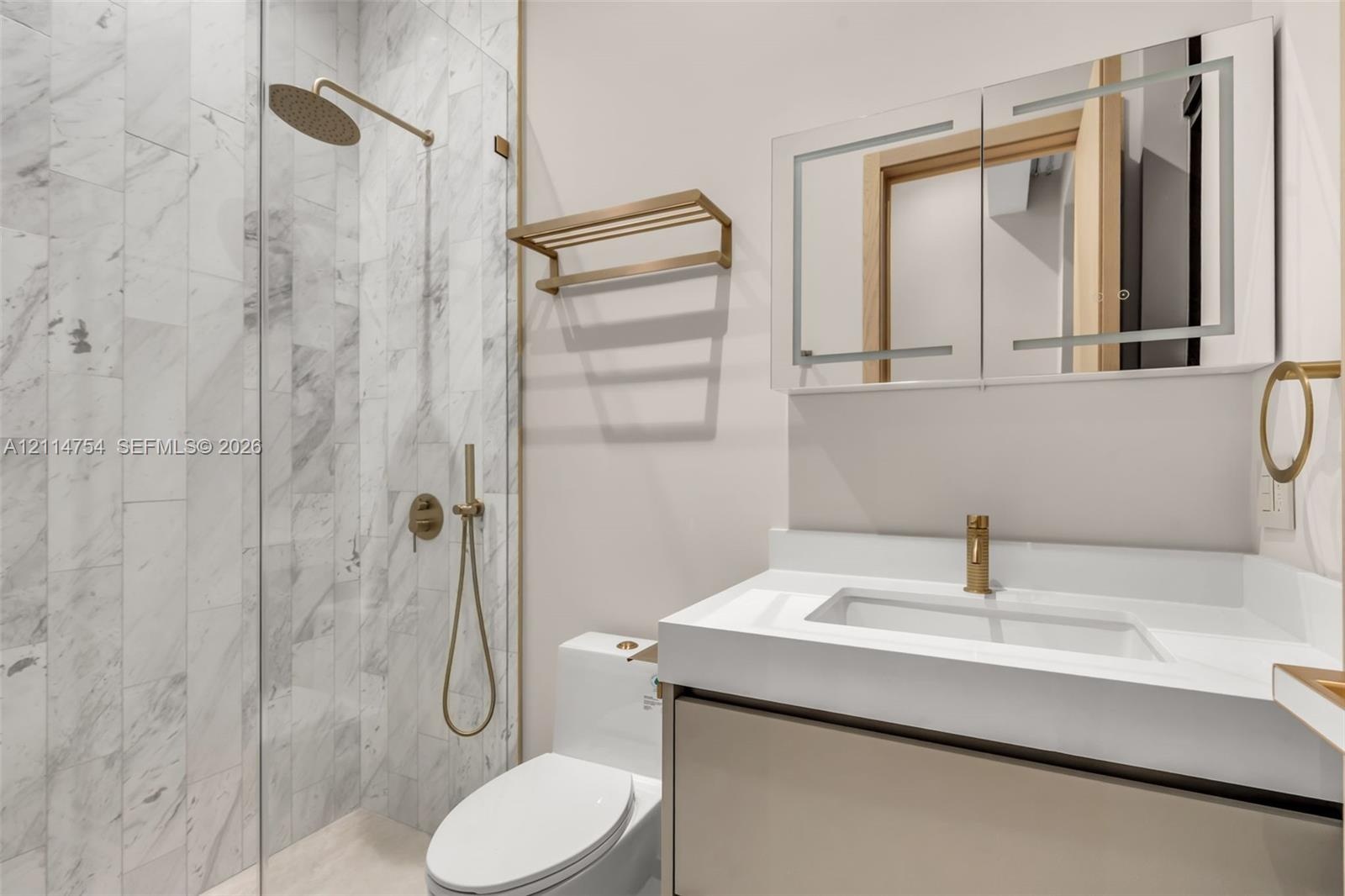 Walk-in shower with vertical stone-finish tiling, brass-toned rainfall shower head, wall-mounted vanity with integrated sink, and illuminated mirrored
