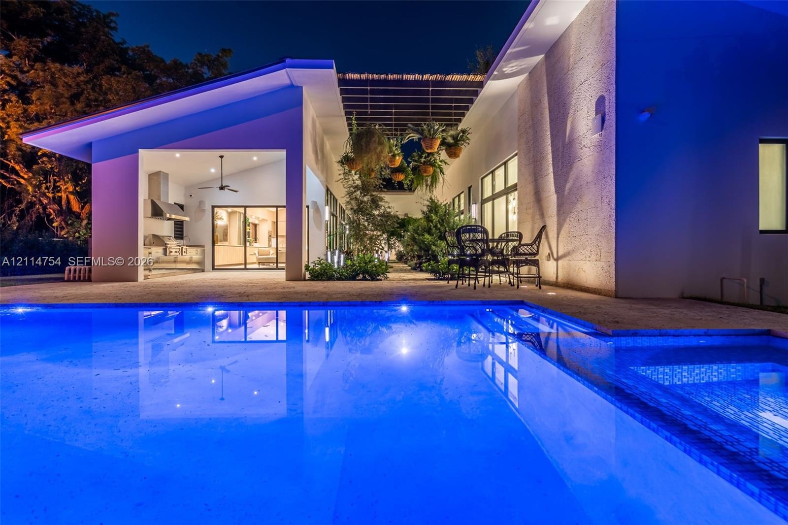 Modern swimming pool featuring stone deck surround, covered outdoor kitchen with stainless steel appliances, and a wood-slat overhead pergola