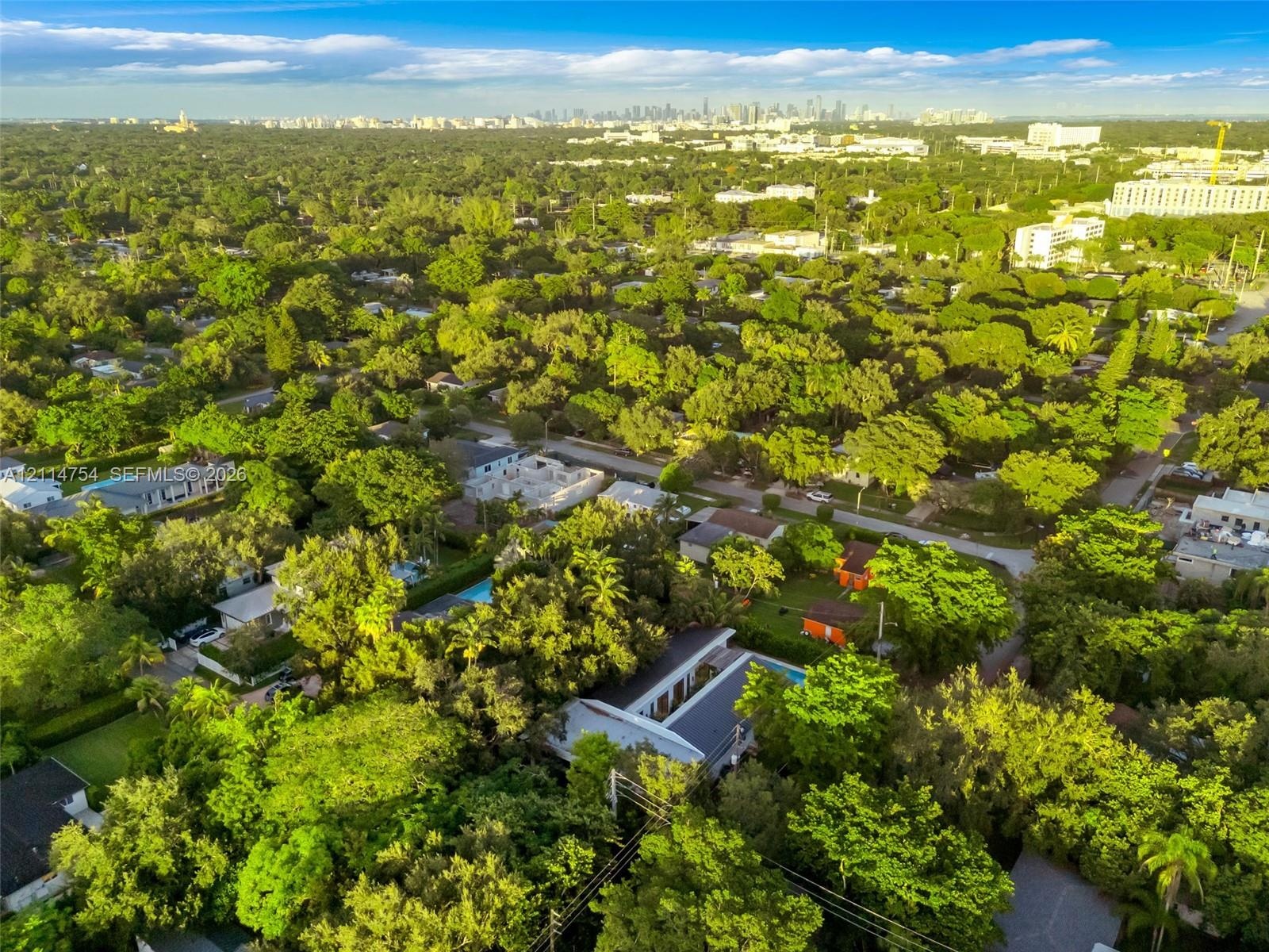 Aerial perspective featuring a flat-roof residence, a rectangular swimming pool, and mature canopy trees, set against a distant cityscape backdrop