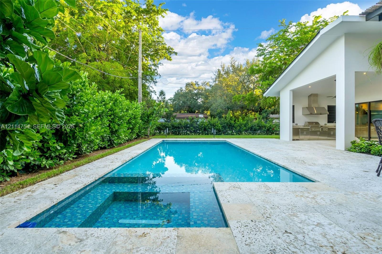 Backyard featuring a rectangular swimming pool with integrated spa, stone-finish decking, covered patio, and built-in outdoor kitchen with stainless s