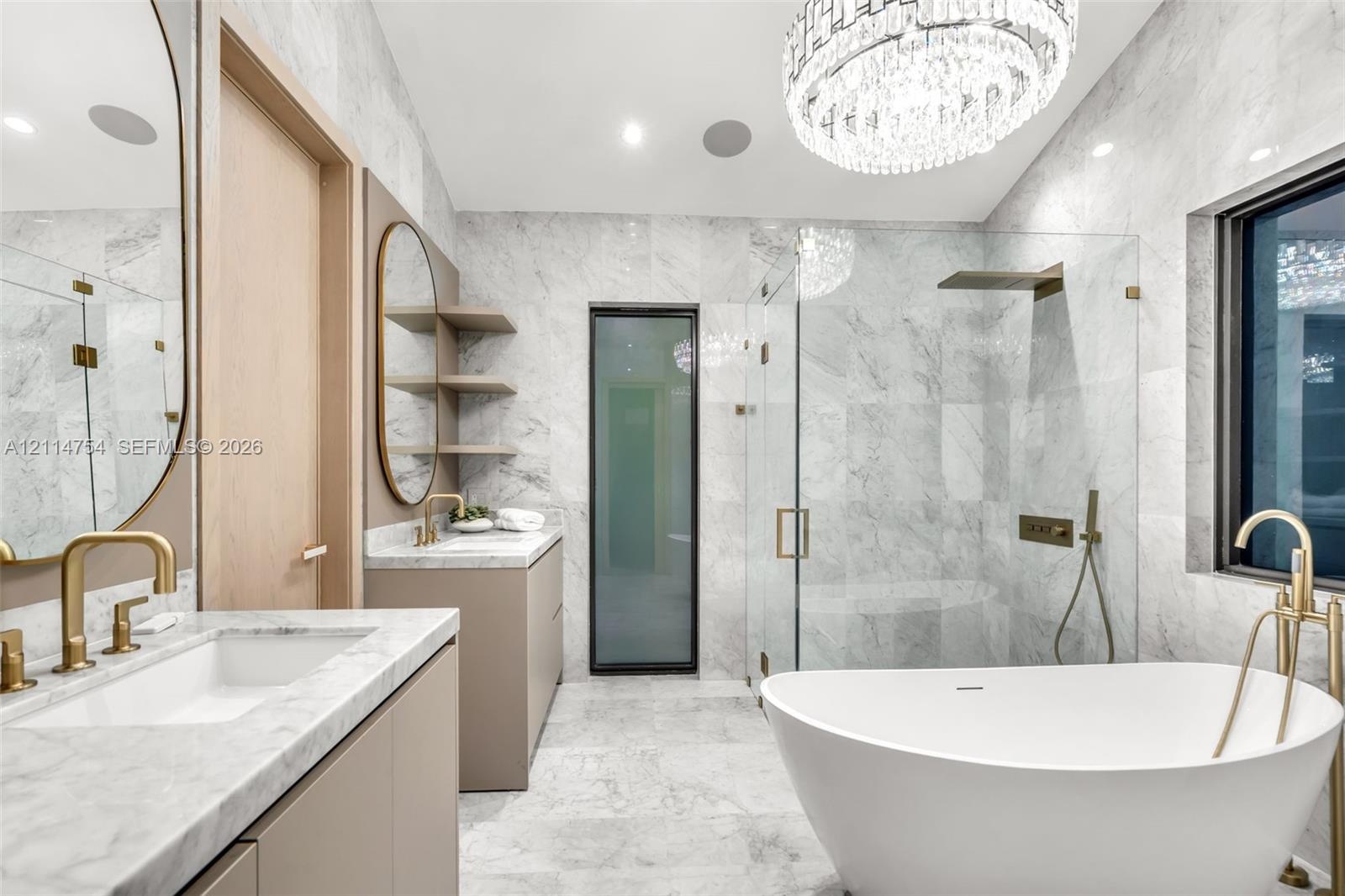 Stone-tiled walls and flooring paired with a freestanding soaking tub, brass fixtures, glass-enclosed walk-in shower, and dual vanities with integrate