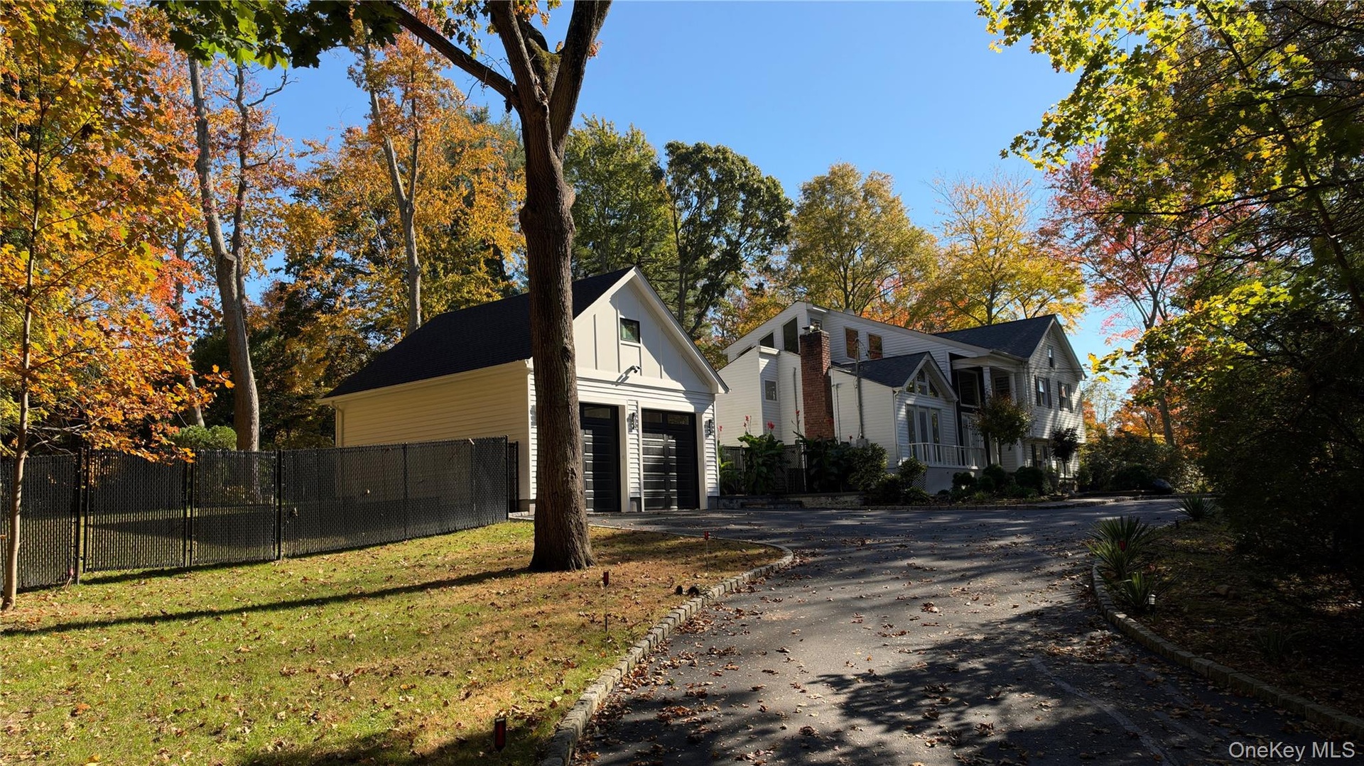 Multi-level residence with white siding and a brick chimney, featuring a detached two-car garage, asphalt driveway, and black chain-link fencing