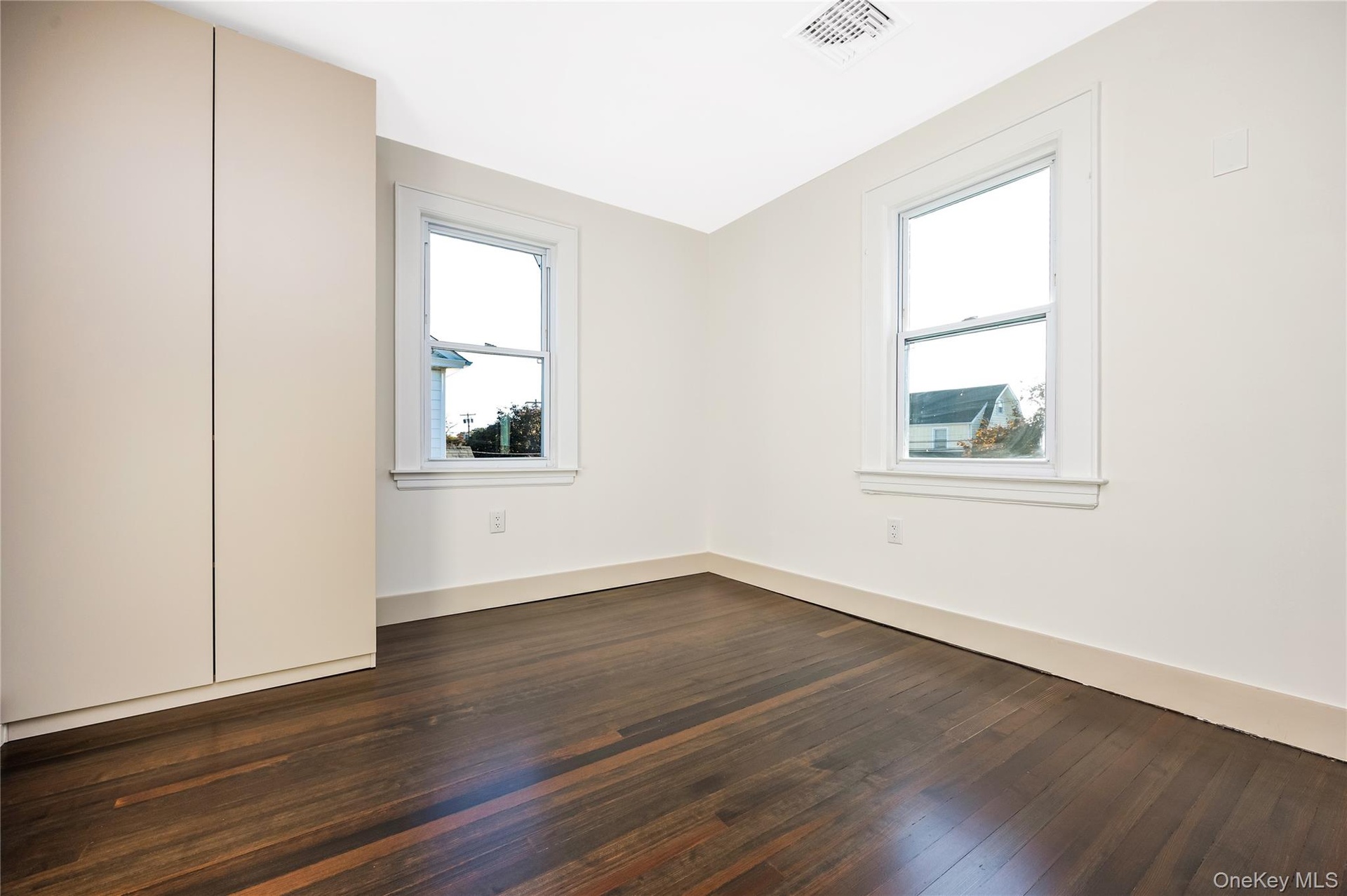 Bedroom featuring dark wood-finish flooring, neutral wall tones, dual windows with white trim, and a floor-to-ceiling built-in storage cabinet