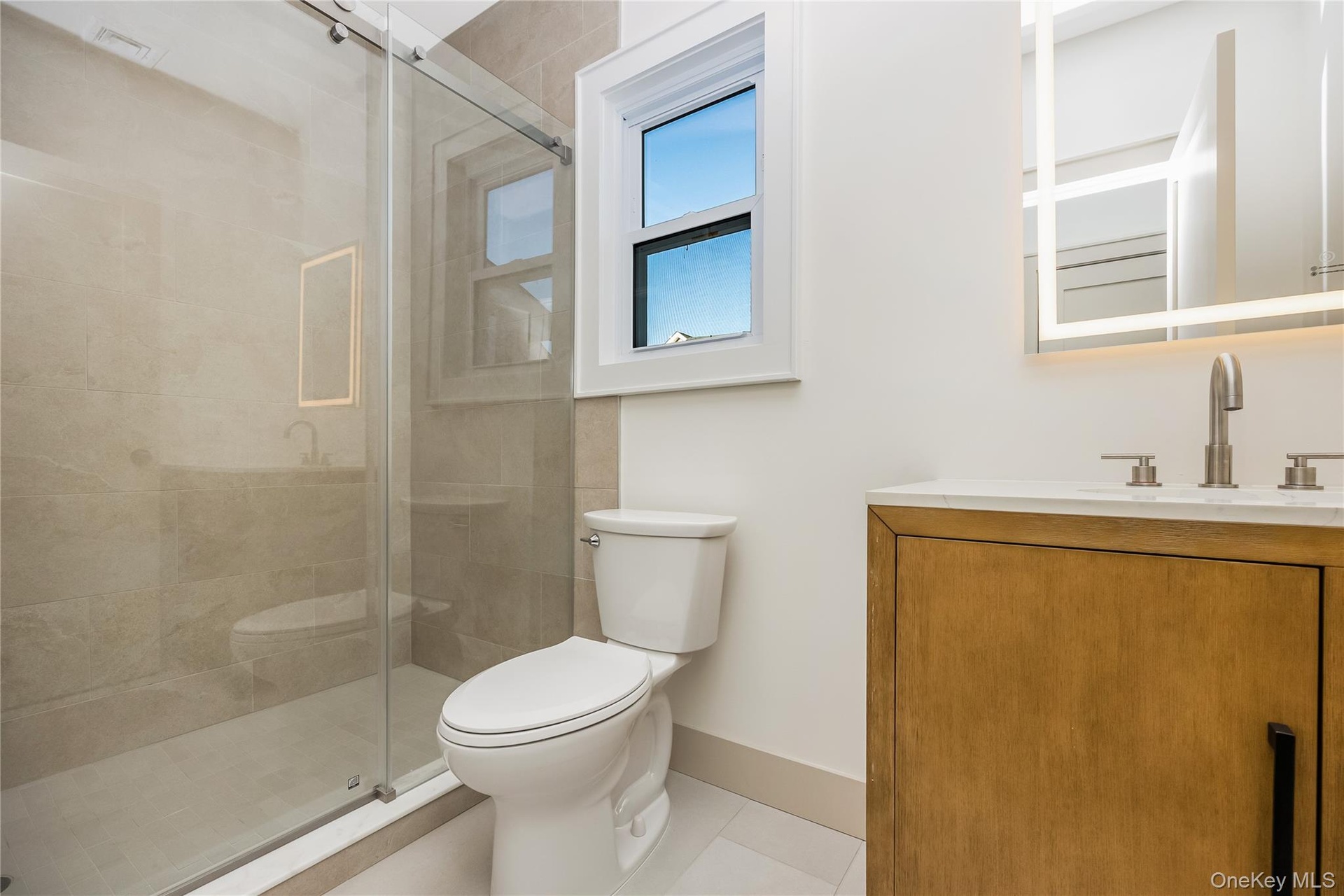 Full bathroom on first floor. Stall shower with glass enclosure and neutral tile surround, vanity with stone-surface countertop, and backlit wall mirr