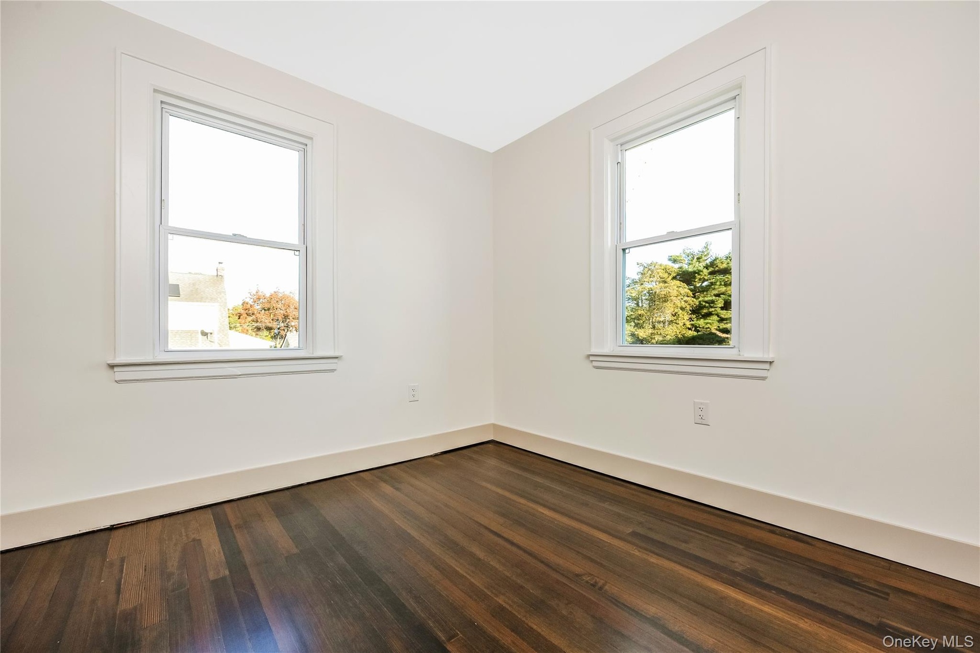 Bedroom with dark-toned wood-finish flooring paired with neutral-colored walls, featuring dual double-hung windows with white trim