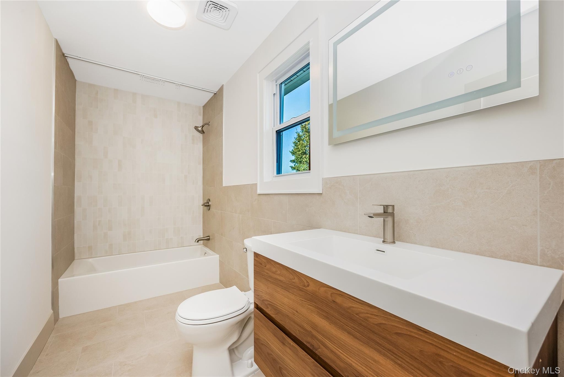 Upstairs full bathroom featuring a floating wood-finish vanity with a rectangular sink, wall-mounted illuminated mirror, neutral wall tiling, and a bu