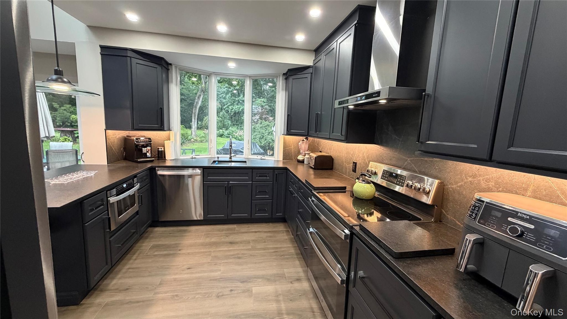 Kitchen featuring dark cabinetry, stone countertops, stainless steel appliances, wood-finish flooring, and a bay window