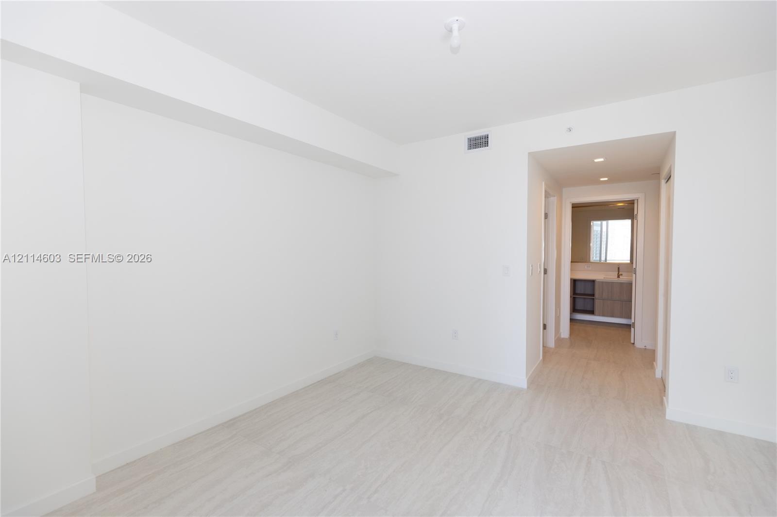 Neutral interior with light-toned wood-finish flooring, smooth white walls, recessed ceiling lighting, and a doorway showcasing a multi-drawer vanity