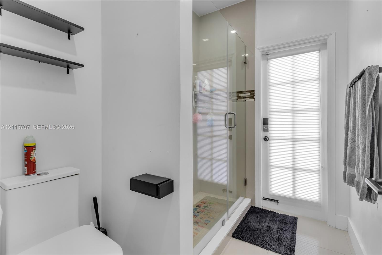 Full-height glass-enclosed shower, wall-mounted matte black shelving, matte black wall-mounted toilet paper holder, and a glass-paneled exterior door