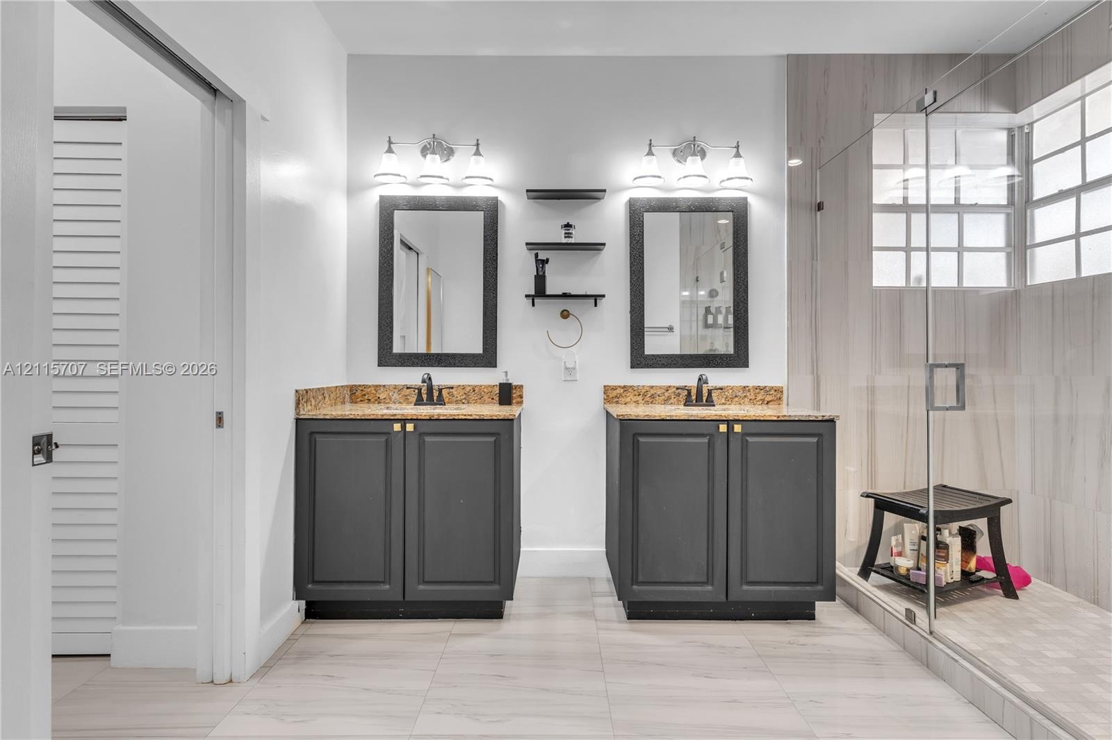Dual-vanity bathroom featuring stone-surfaced countertops, charcoal-finish cabinetry, framed mirrors, and a glass-enclosed shower with tiled wall surr