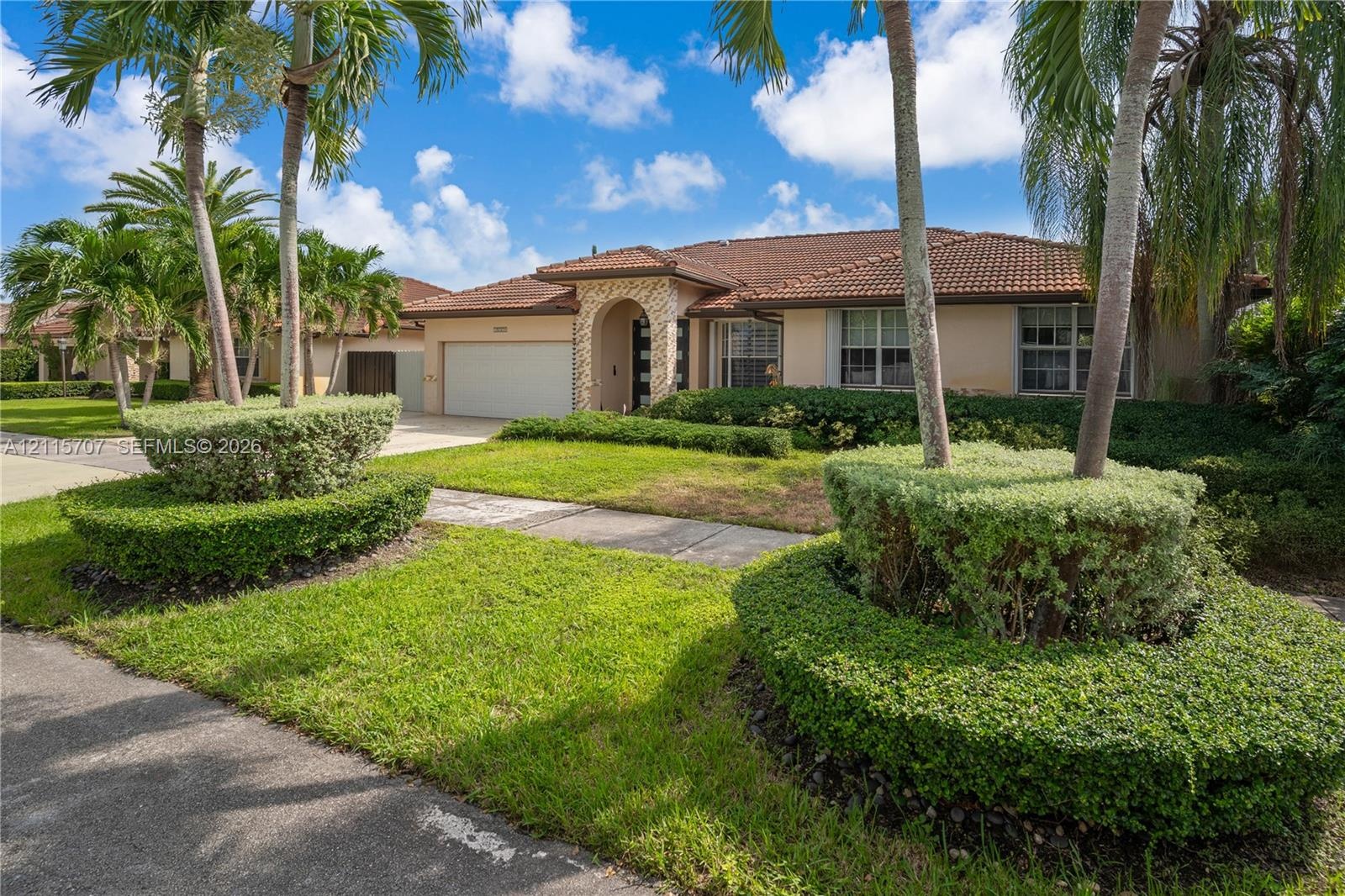 Single-story exterior featuring a concrete tile roof, stone veneer entryway, and two-car garage, surrounded by manicured landscaping and mature palm t