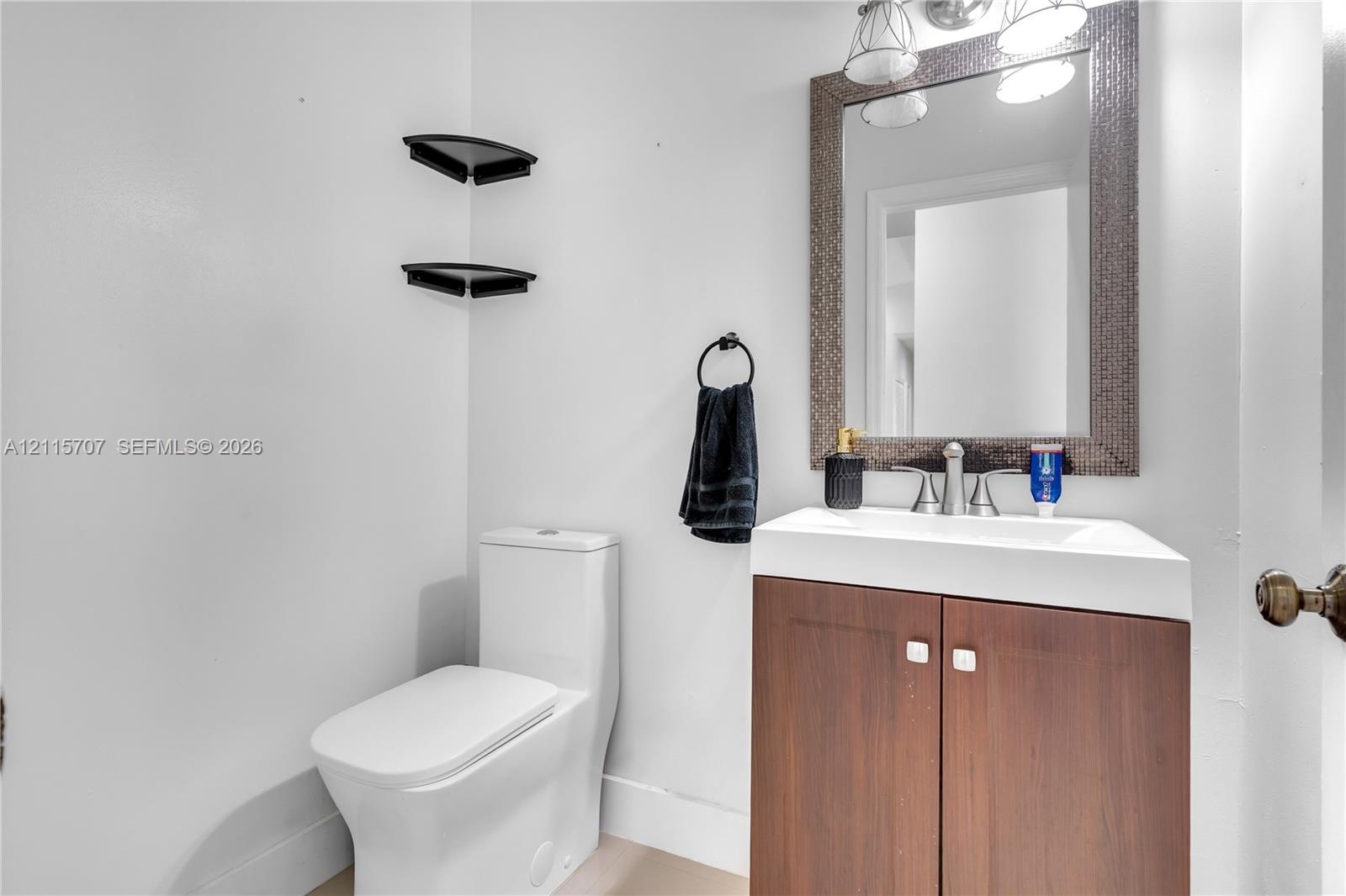 Bathroom featuring a wood-finish vanity with a rectangular sink, wall-mounted corner shelving, a framed mirror with integrated lighting, and a modern 