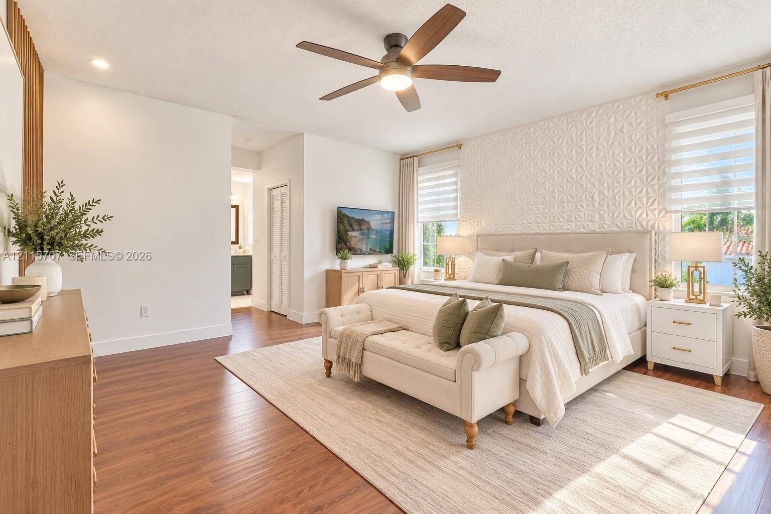 Bedroom featuring wood-finish flooring, a textured geometric accent wall, a central ceiling fan with integrated lighting, and dual windows with layere