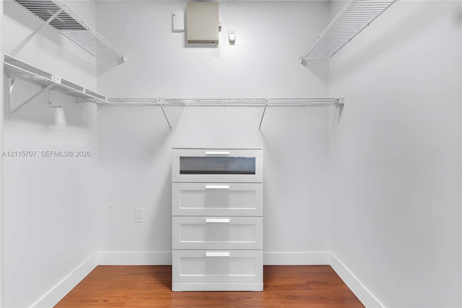 Walk-in closet featuring wood-finish flooring, white built-in four-drawer dresser, and tiered wire shelving