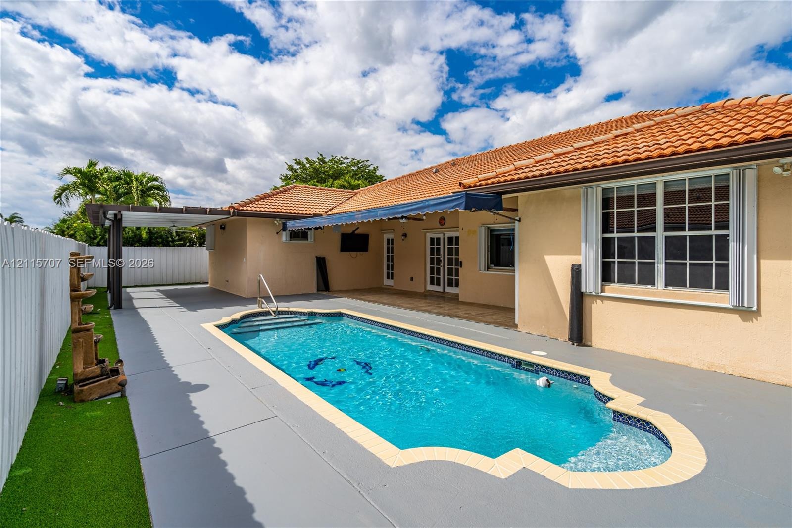 In-ground swimming pool with a custom-shaped perimeter, terra cotta tile roof, retractable fabric awning, wooden-frame pergola, and synthetic turf law