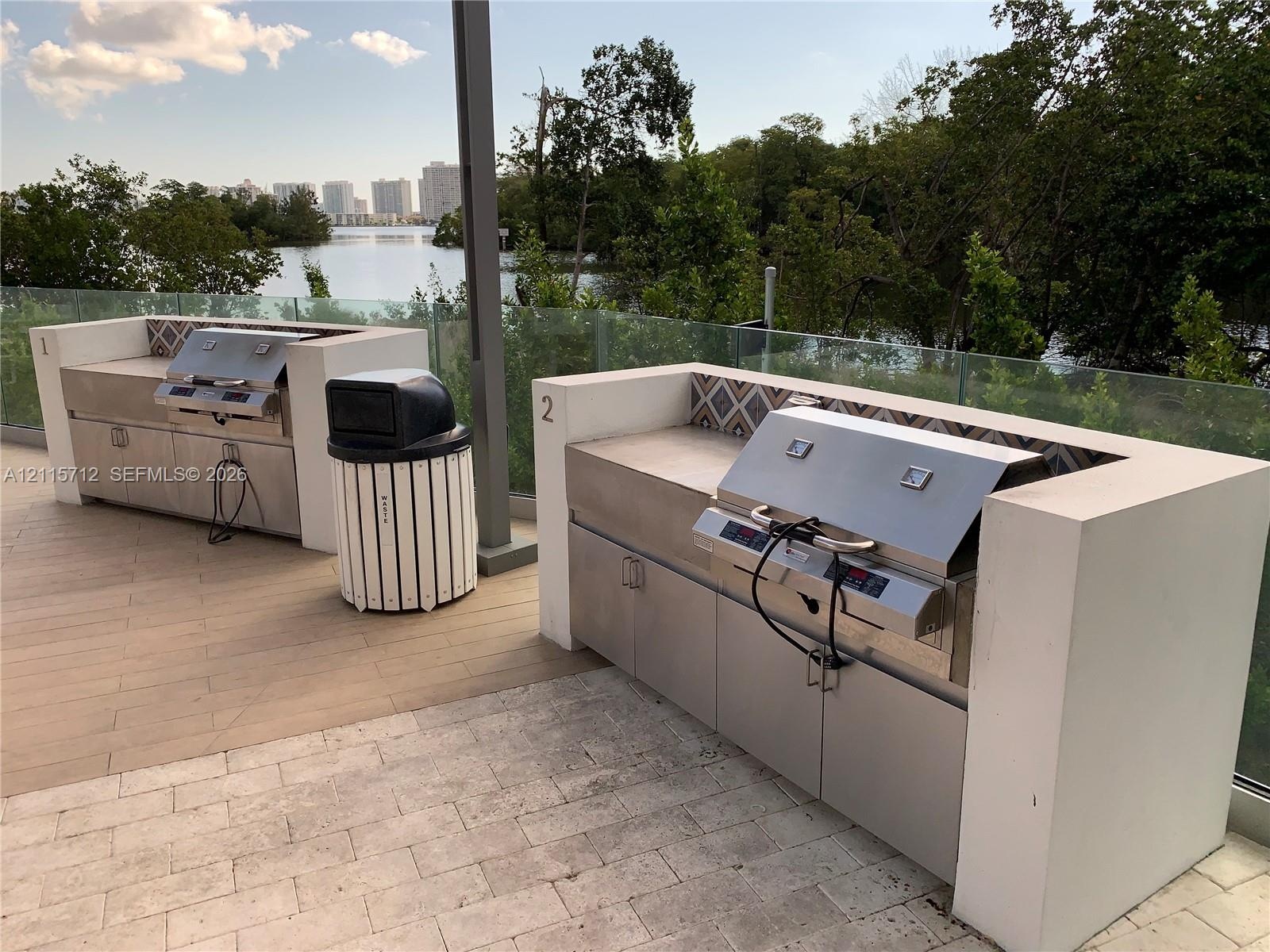 Outdoor kitchen featuring dual stainless steel built-in grills, concrete-surround masonry counters, decorative tile backsplashes, and glass panel rail