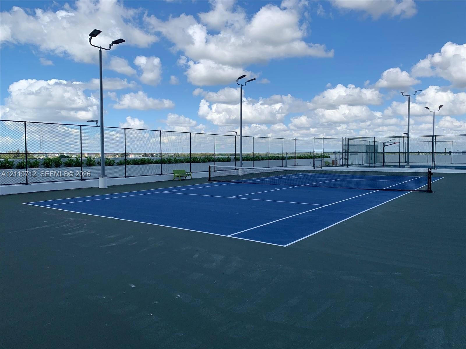 Rooftop tennis court featuring a blue surface with white court markings, perimeter chain-link fencing, and integrated pole-mounted floodlighting