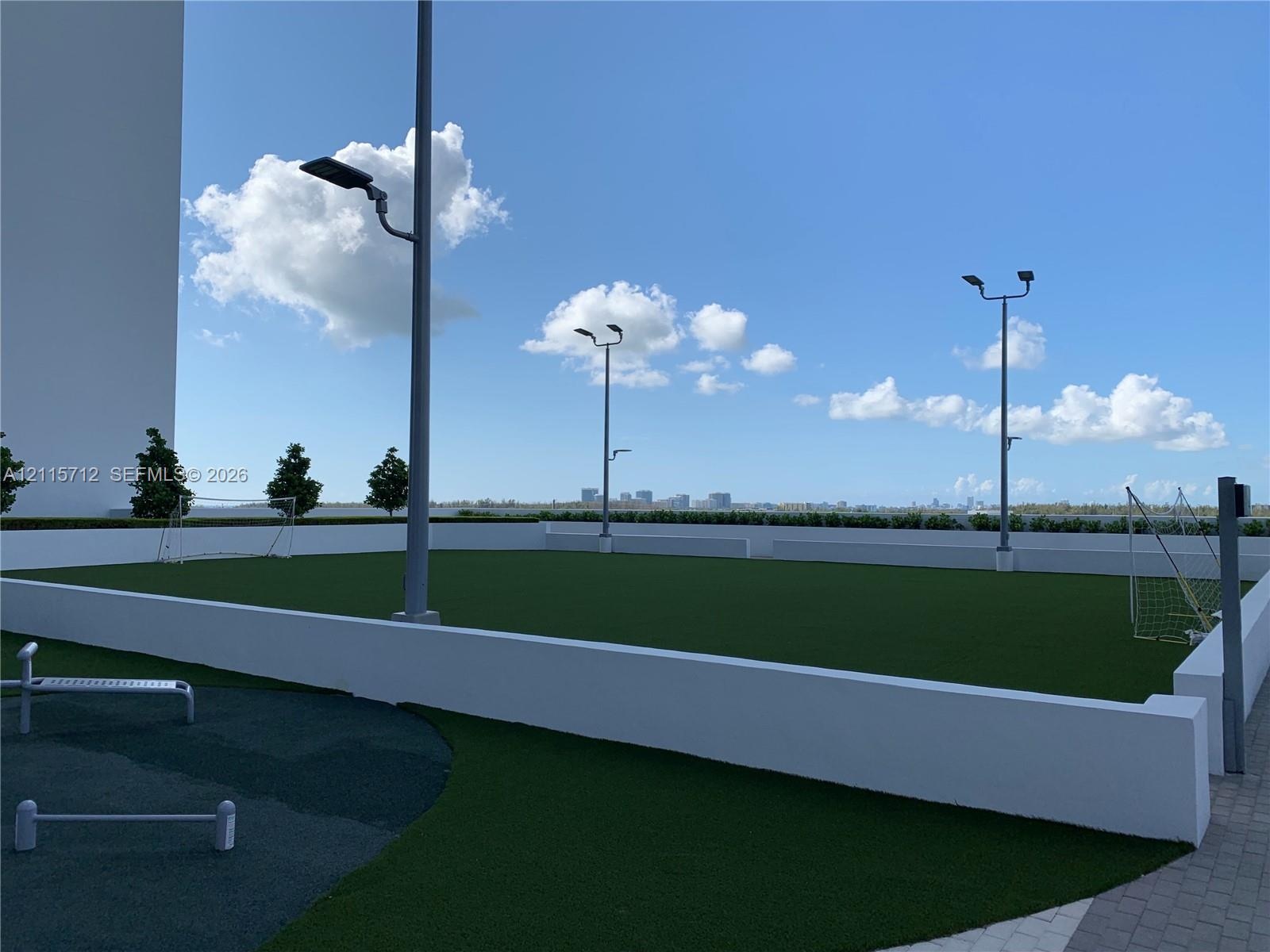 Outdoor recreation area featuring artificial turf, perimeter concrete walls, tall stadium-style lighting, and paved pathways