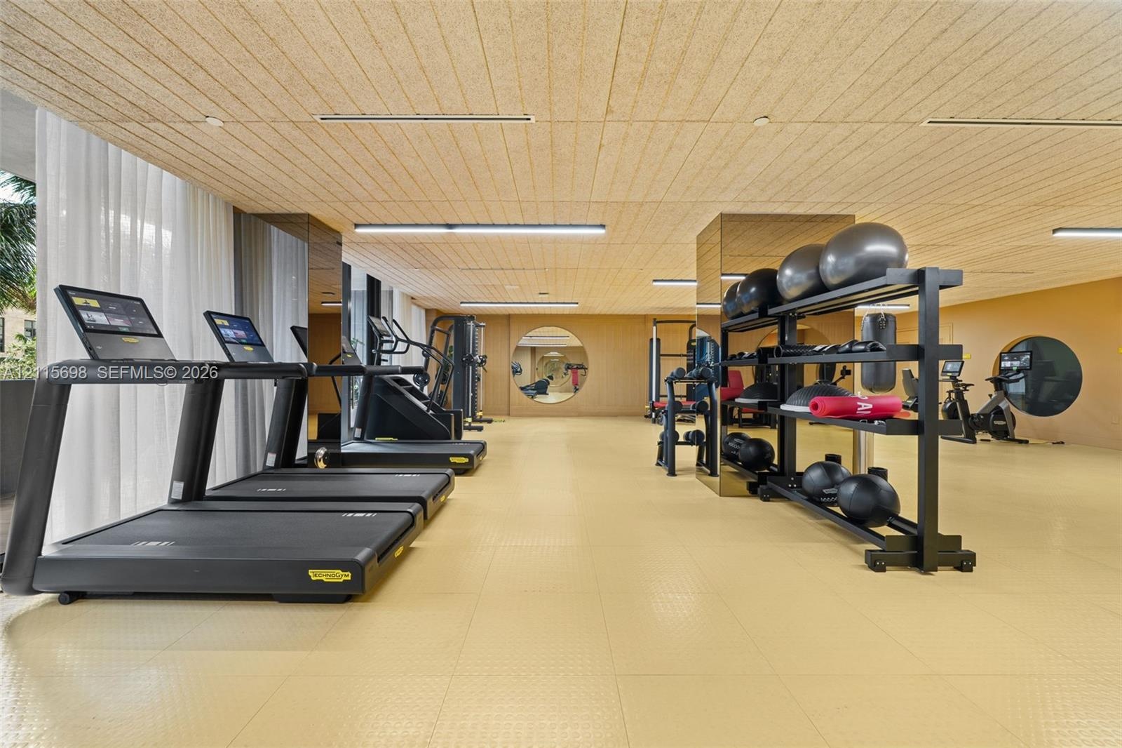 Fitness area featuring textured wood-panel ceiling, mirrored structural pillars, circular accent mirrors, recessed linear lighting, and resilient athl