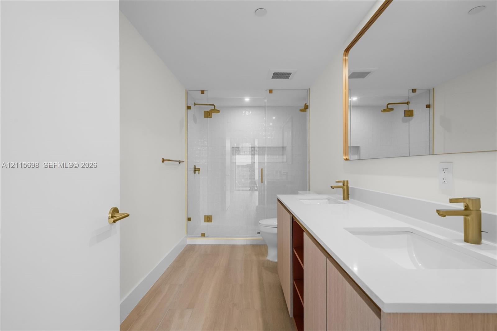Dual-sink vanity with quartz countertop, light wood-finish cabinetry, brass-toned fixtures, rectangular mirror with metallic frame, and a glass-enclos