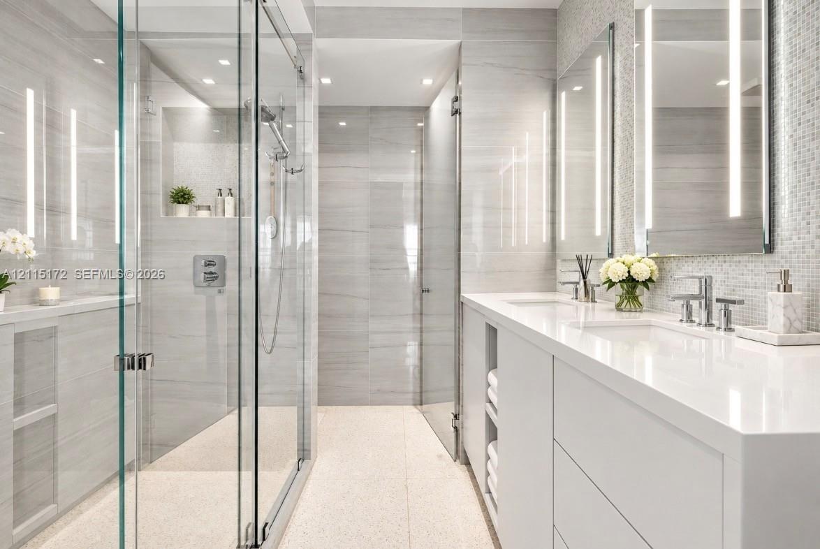 Walk-in glass-enclosed shower featuring large-format tile wall cladding, integrated wall niche, dual-sink vanity with white stone countertop, and vert