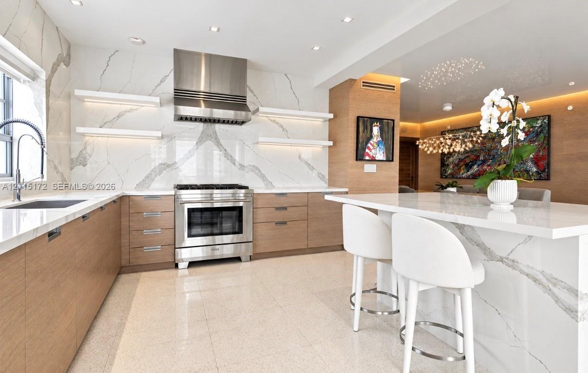 Kitchen featuring flat-panel wood cabinetry, a waterfall-edge island, a floor-to-ceiling stone-slab backsplash, integrated stainless steel range, and 