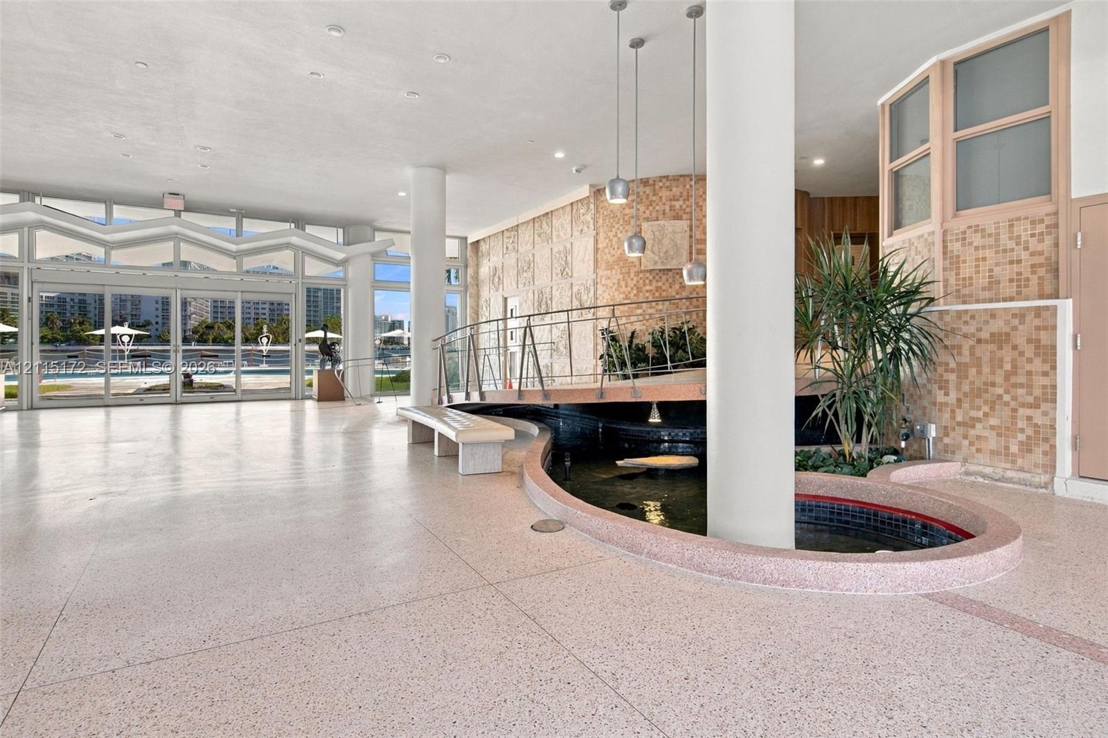 Spacious interior featuring terrazzo flooring, a central decorative pond, full-height glass folding doors, structural cylindrical columns, and pendant