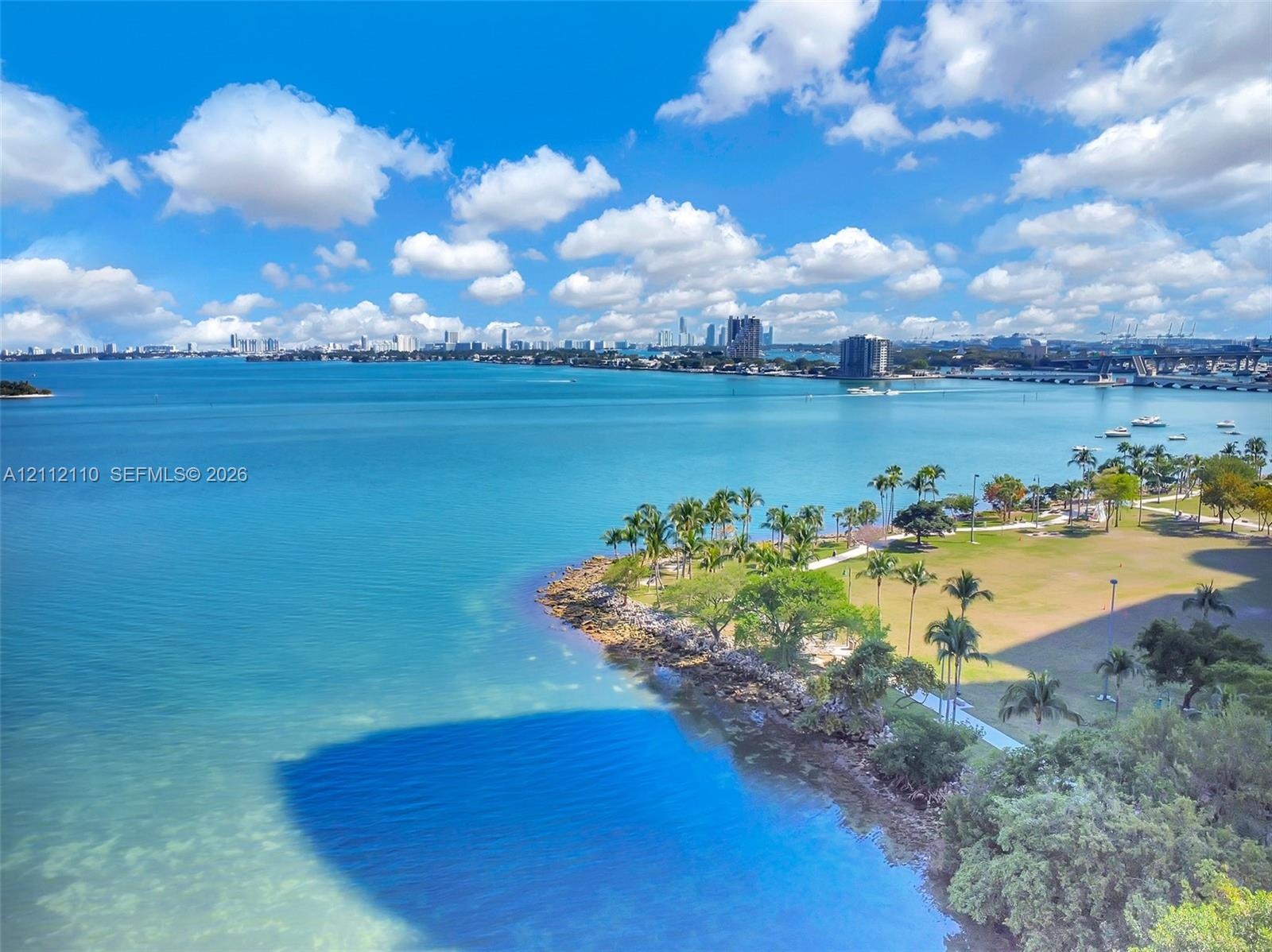 Panoramic waterfront scenery featuring open bay waters, coastal shoreline, manicured green space, palm trees, and a skyline horizon