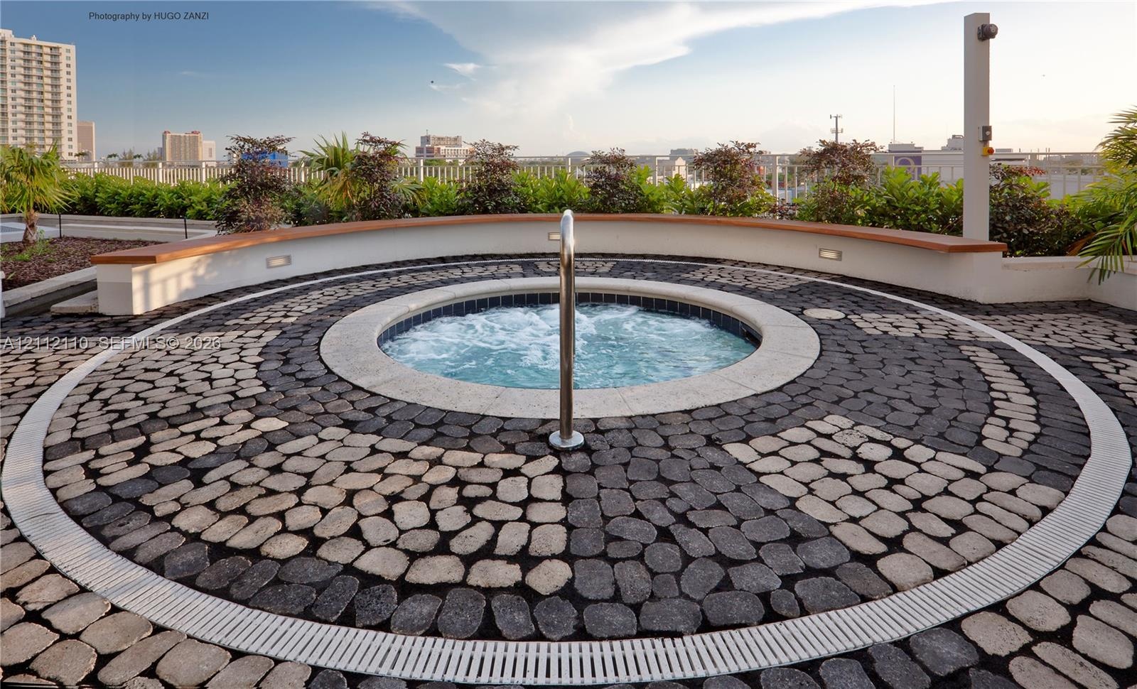 Circular in-ground spa featuring a stone-tiled deck, a smooth masonry surround, integrated perimeter lighting, and metallic handrail