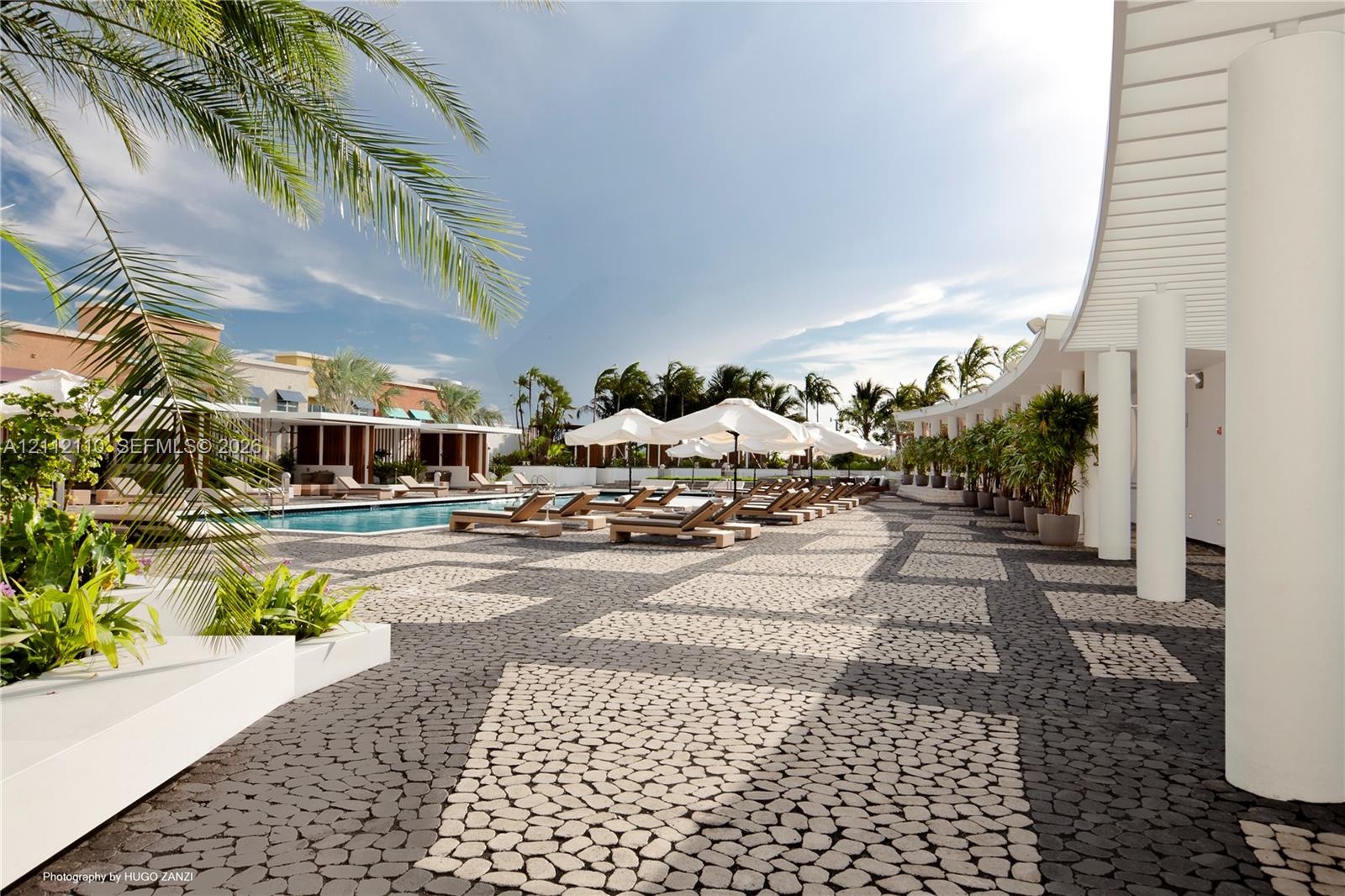 Expansive swimming pool area featuring a curved architectural overhang with structural columns, patterned stone masonry decking, and integrated plante