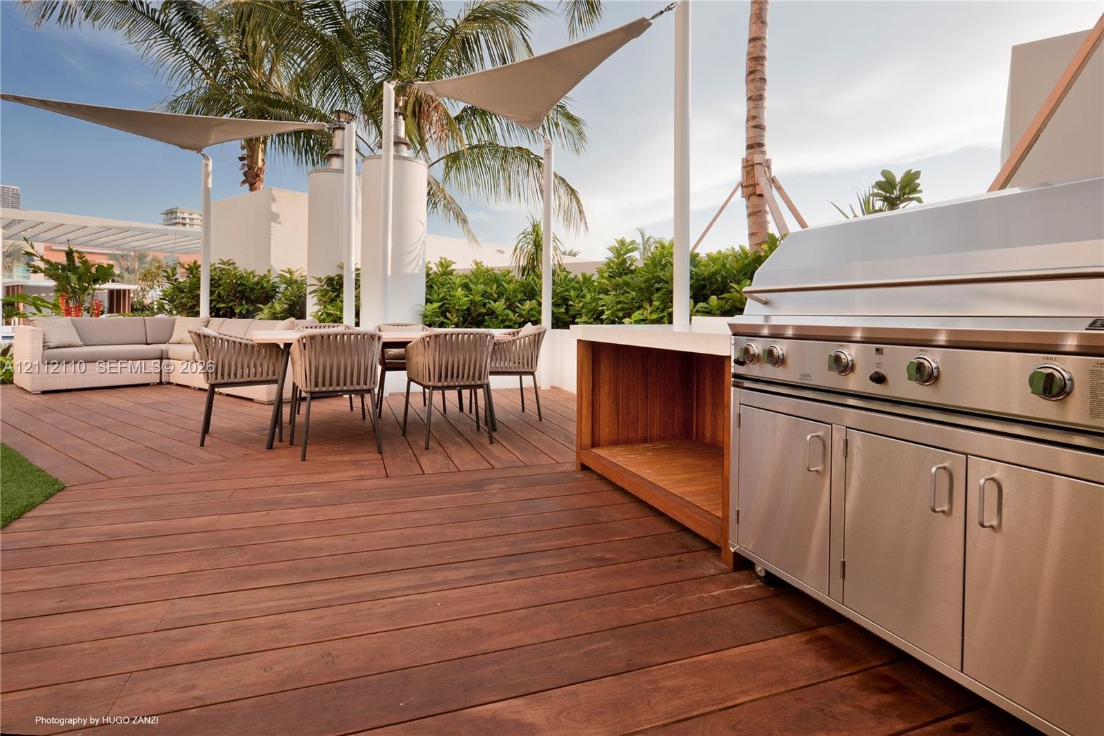 Outdoor terrace featuring wood-finish decking, a built-in stainless steel grill station, white structural shade sails, and integrated landscaping