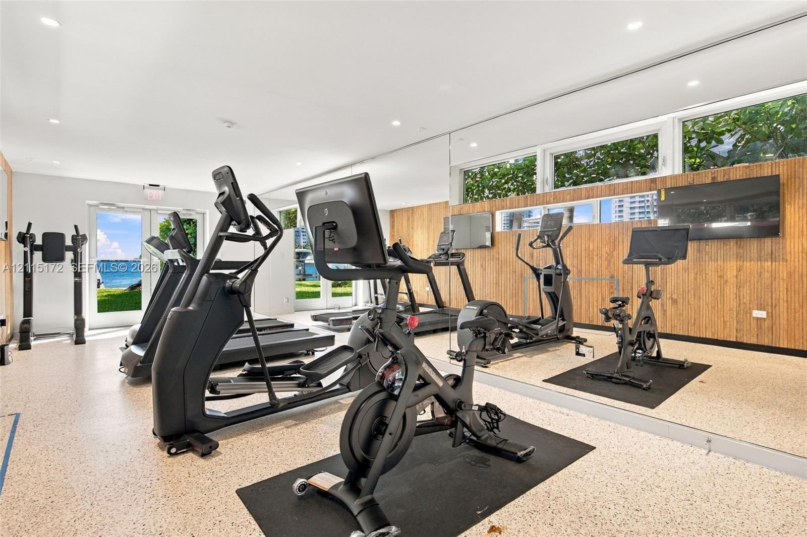 Fitness area featuring full-wall mirrors, recessed lighting, wood-slat accent wall cladding, and floor-to-ceiling glass doors providing natural light