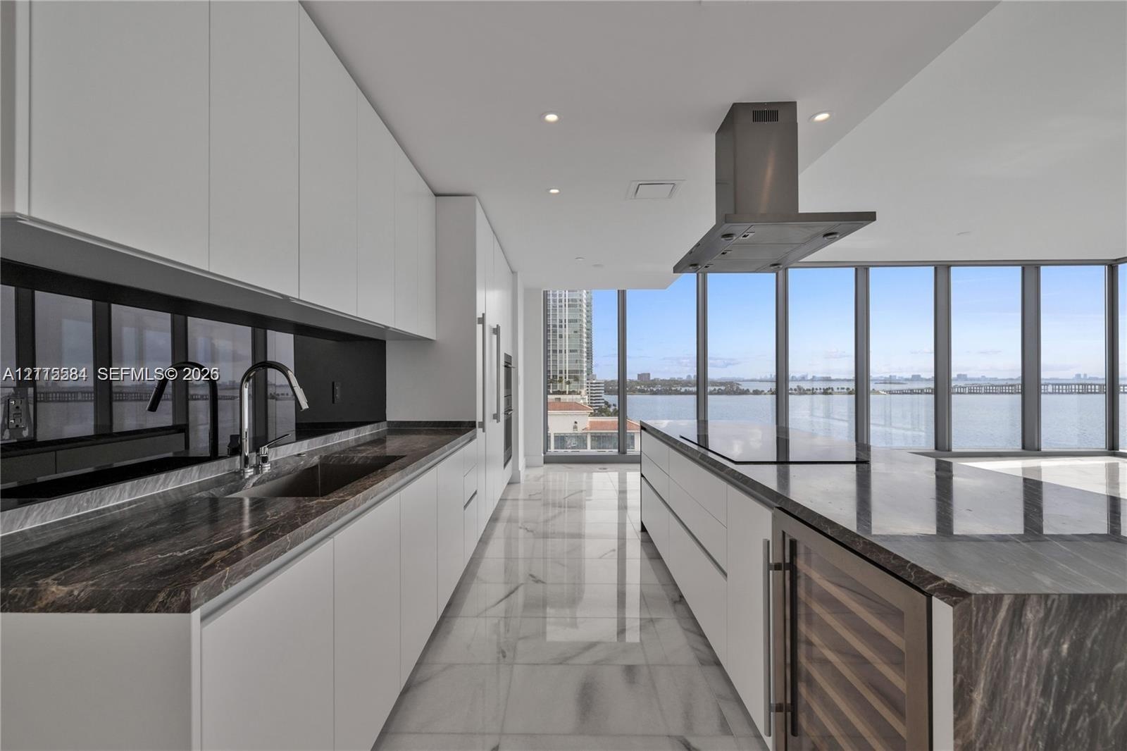 Contemporary kitchen featuring white flat-panel cabinetry, stone countertops, integrated wine refrigeration, stainless steel vent hood, and floor-to-c