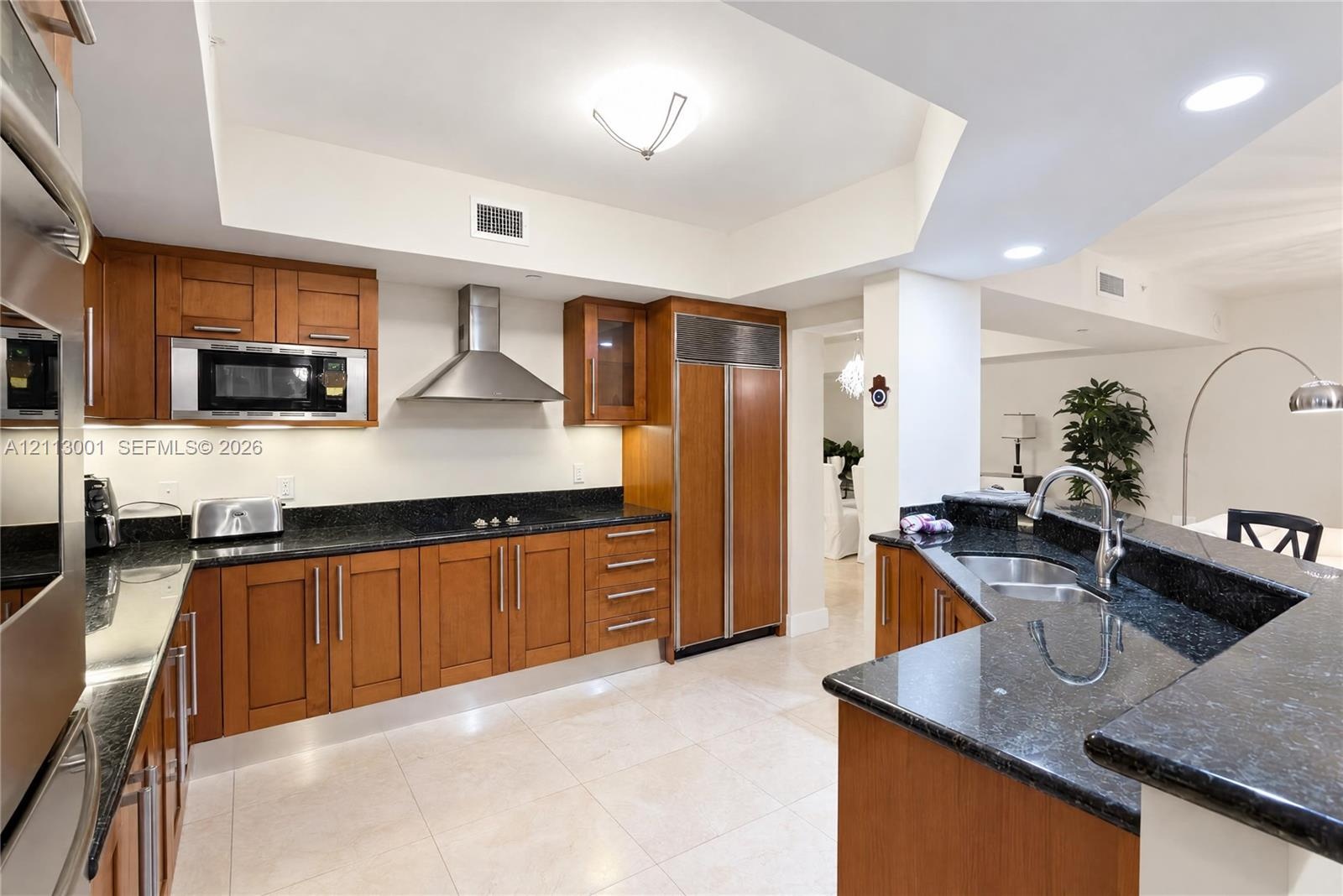Kitchen featuring wood cabinetry, dark stone countertops, stainless steel appliances including a built-in refrigerator and wall oven, recessed lightin