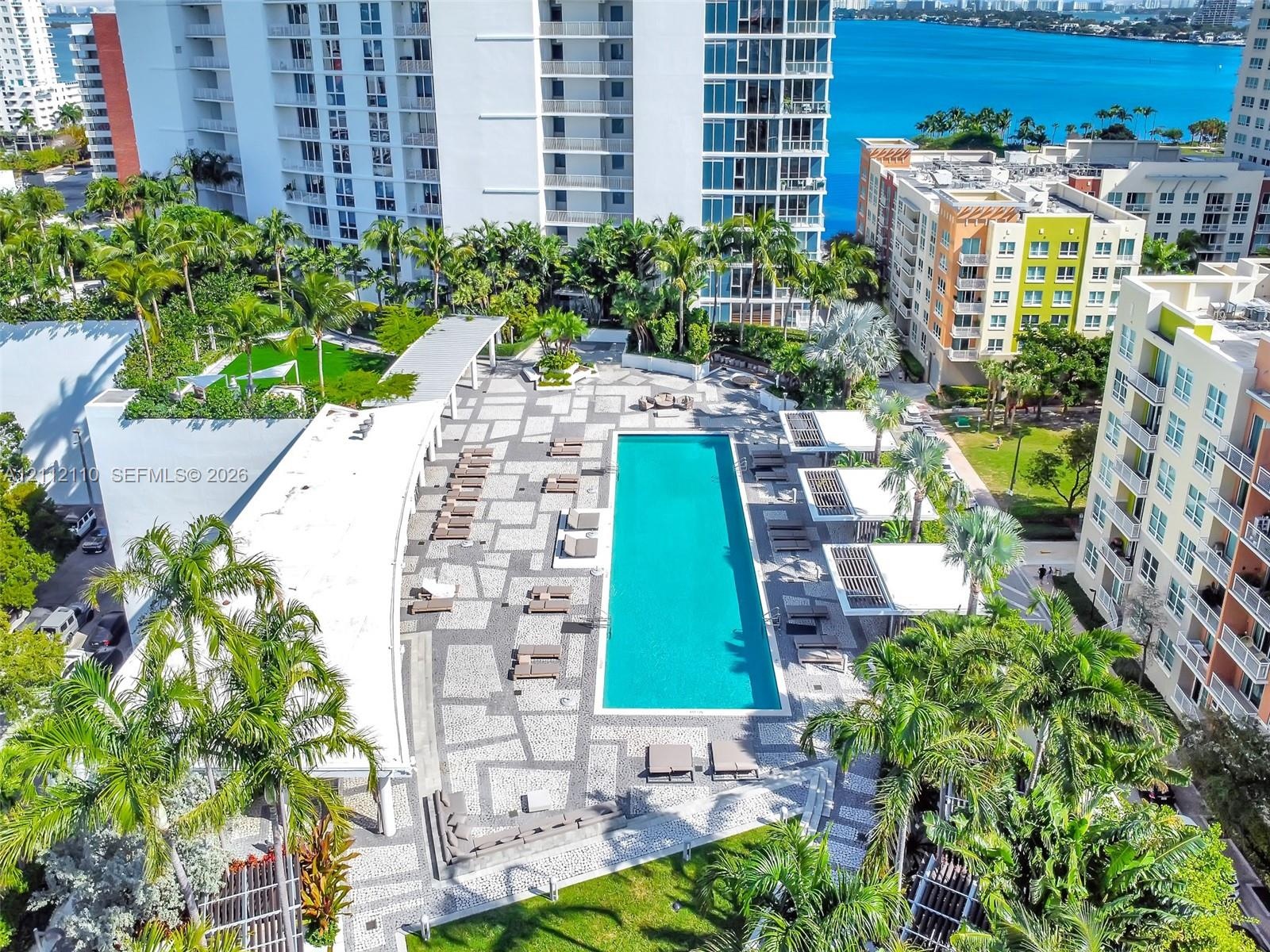 Rectangular swimming pool featuring a geometric paver deck, covered patio structures, and perimeter landscaping with mature palms