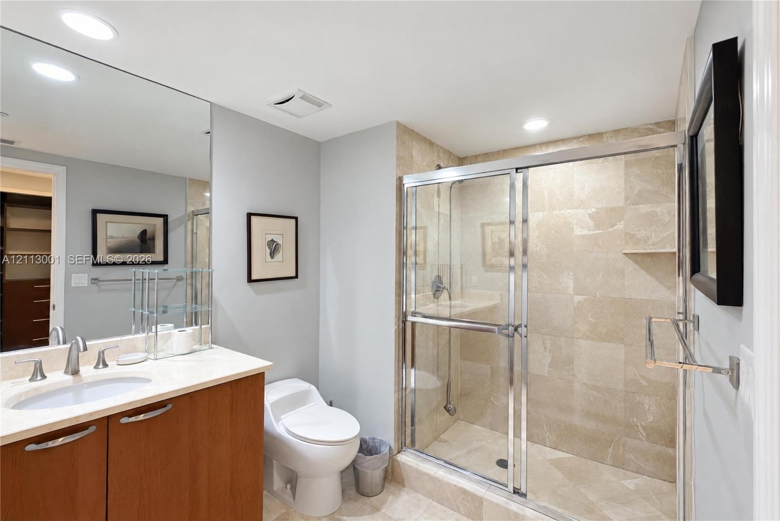 Bathroom featuring a wood-finish vanity with a light-toned countertop, integrated oval sink, full-width wall mirror, and walk-in shower with tiled wal