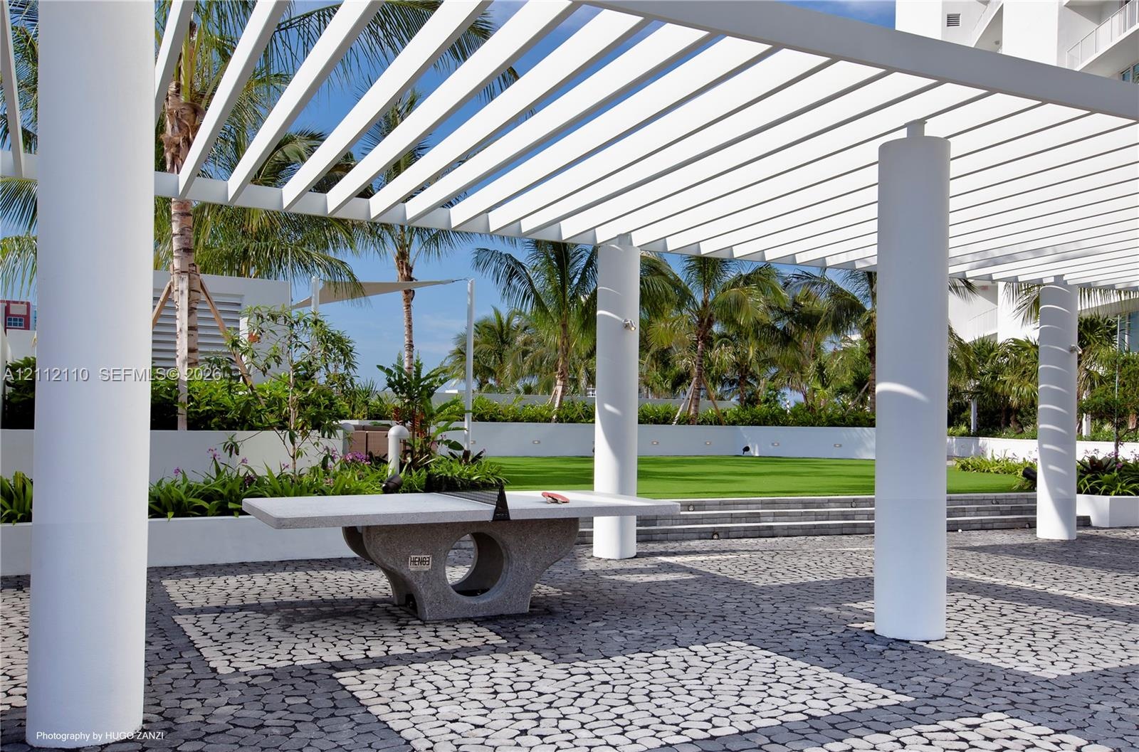 Outdoor space featuring a white slatted pergola, cylindrical support columns, stone paver flooring, a permanent stone table, and a tiered lawn area