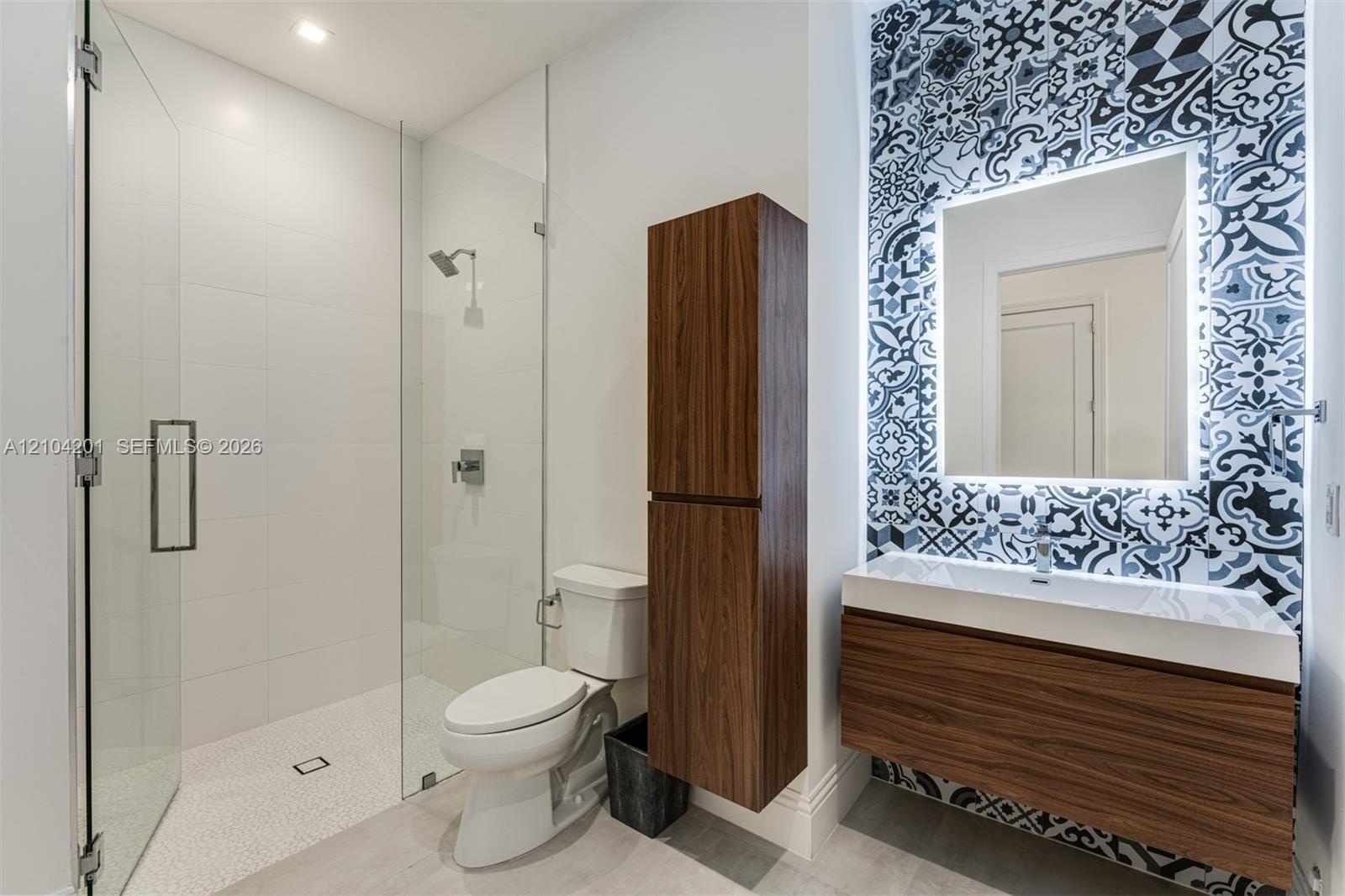Bathroom featuring a walk-in shower with a frameless glass enclosure, wall-mounted wood-finish vanity, illuminated mirror, decorative patterned wall t