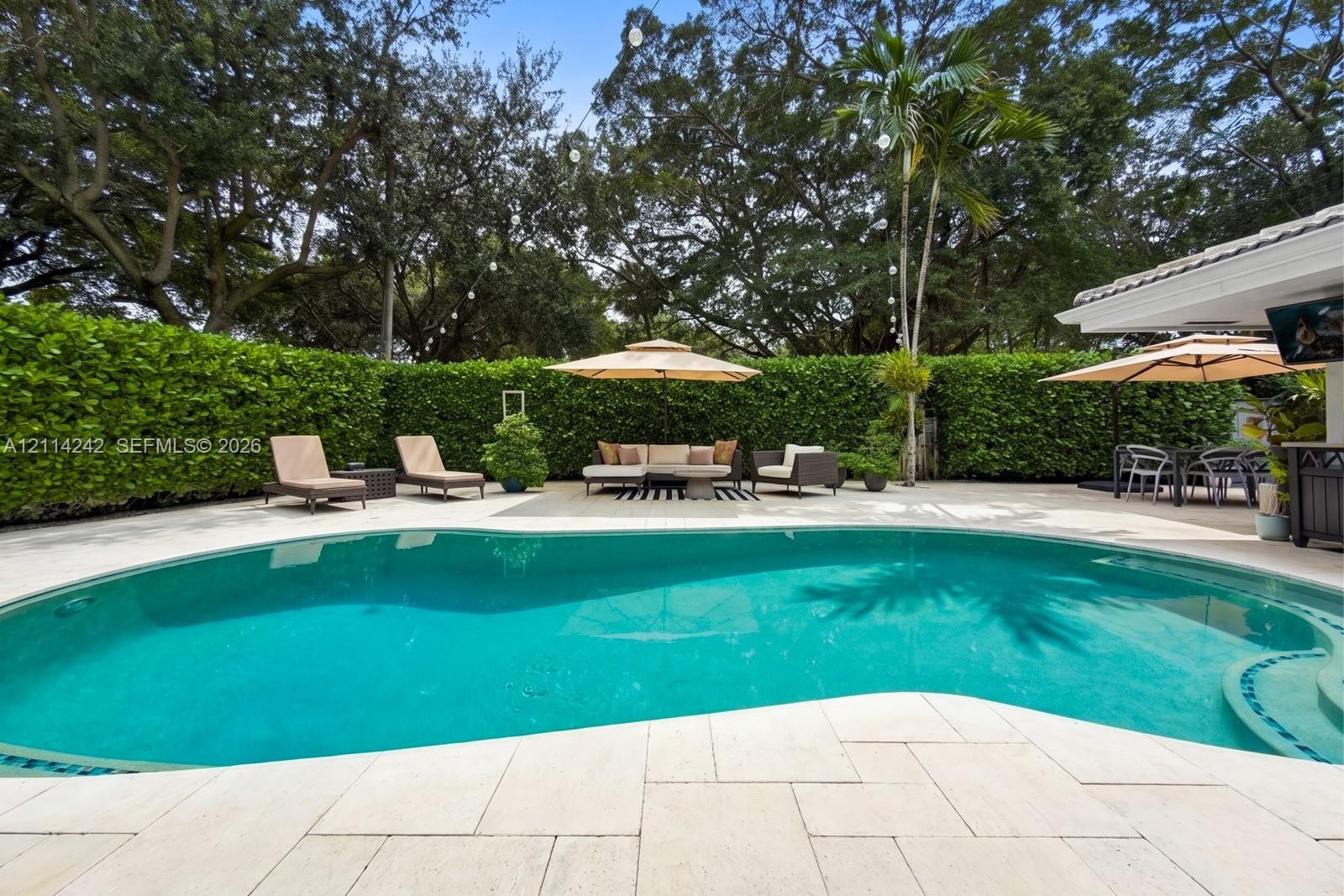 Curved swimming pool with built-in stairs, large-format stone paver decking, and extensive mature privacy hedging