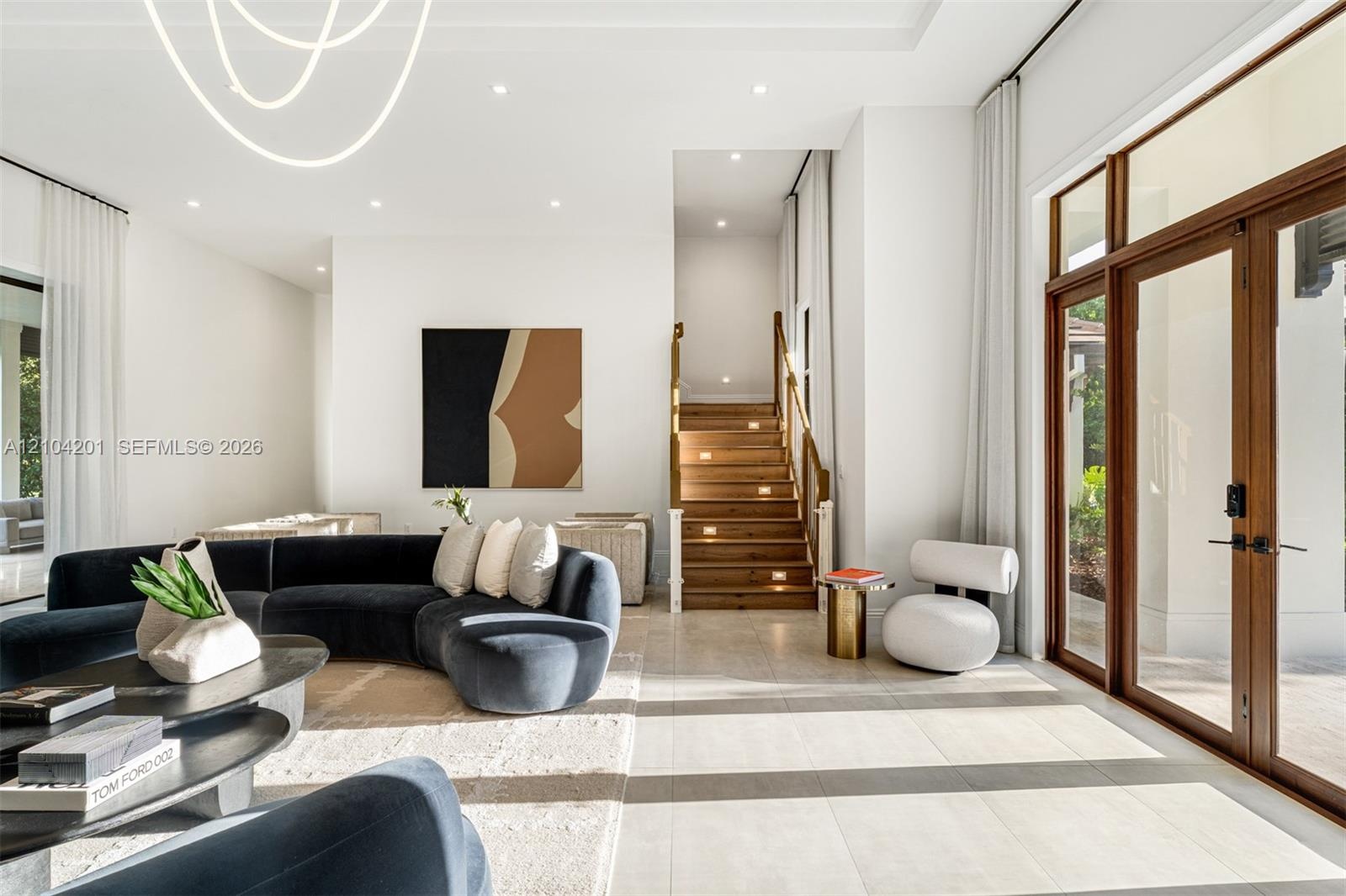 High-ceiling living area featuring large-format tile flooring, double glass doors with wood-finish frames, a recessed tray ceiling, and a staircase wi