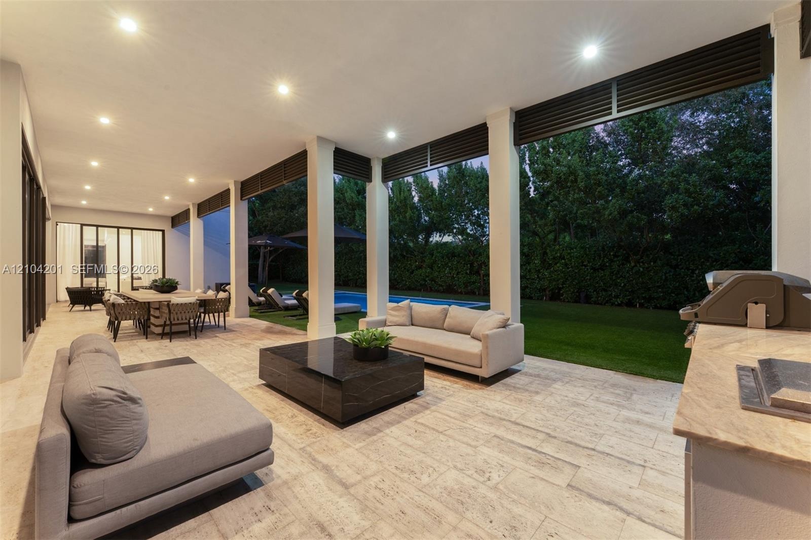Covered outdoor living area featuring light-toned stone flooring, recessed ceiling lighting, structural support columns, an integrated outdoor kitchen