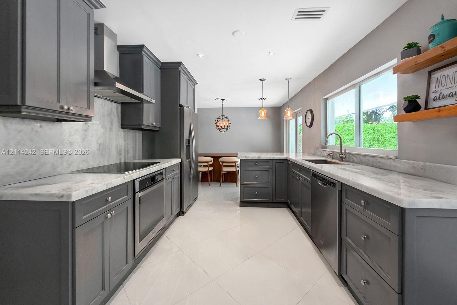 Kitchen featuring charcoal-toned cabinetry, light-toned stone-finish countertops, full-height stone-finish backsplash, stainless steel appliances, and