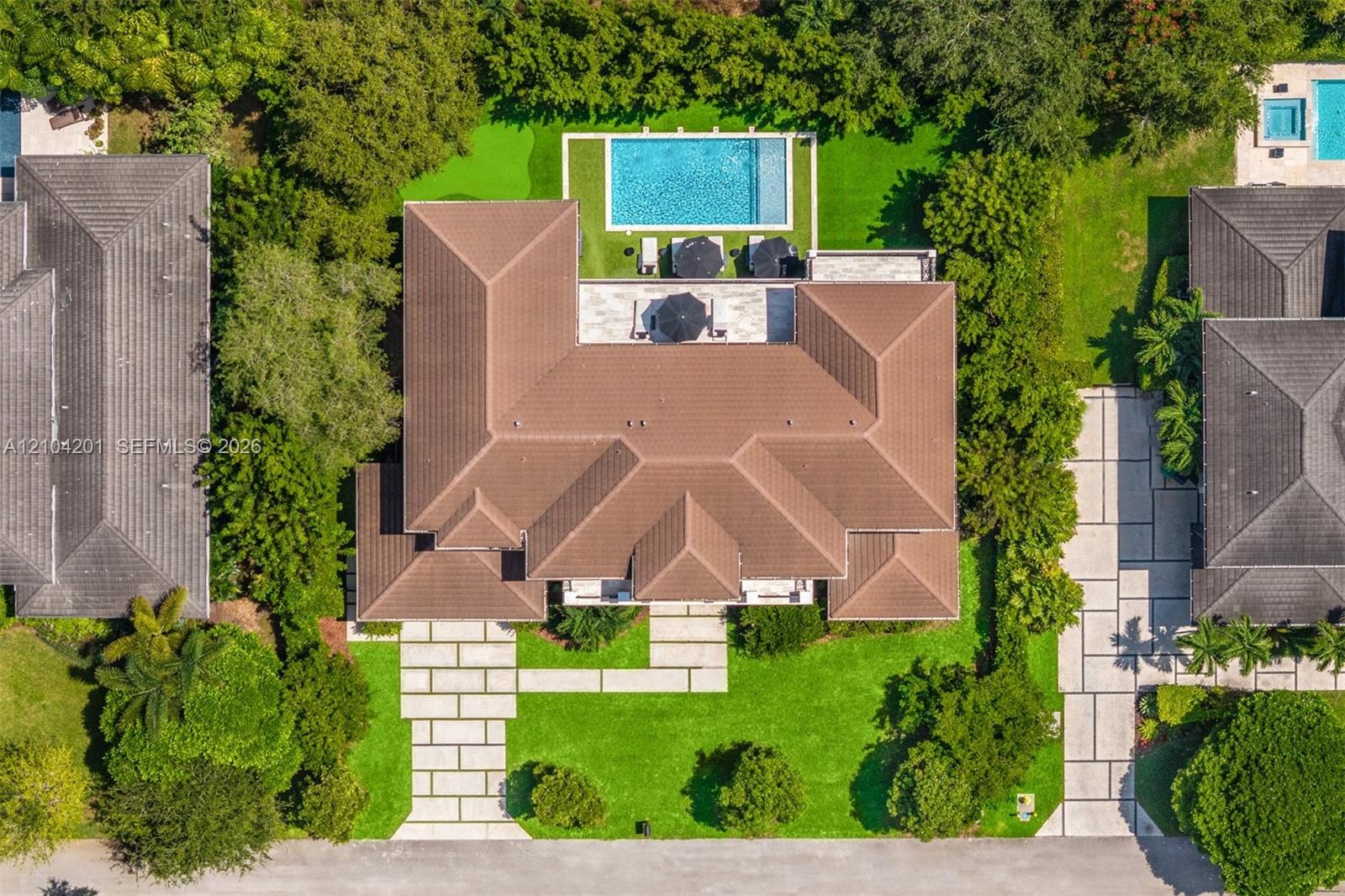 Aerial perspective of a property featuring a tiled roof, rectangular swimming pool, stone paver driveway, manicured lawn, and mature perimeter landsca
