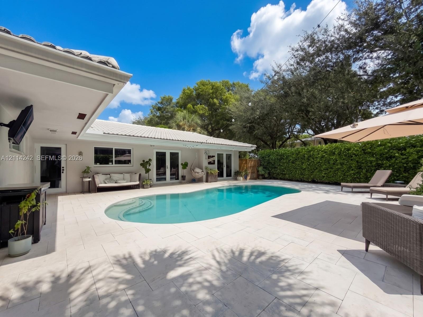 Backyard oasis featuring an in-ground pool with integrated steps, expansive stone-paver patio, covered porch area, and natural hedge perimeter