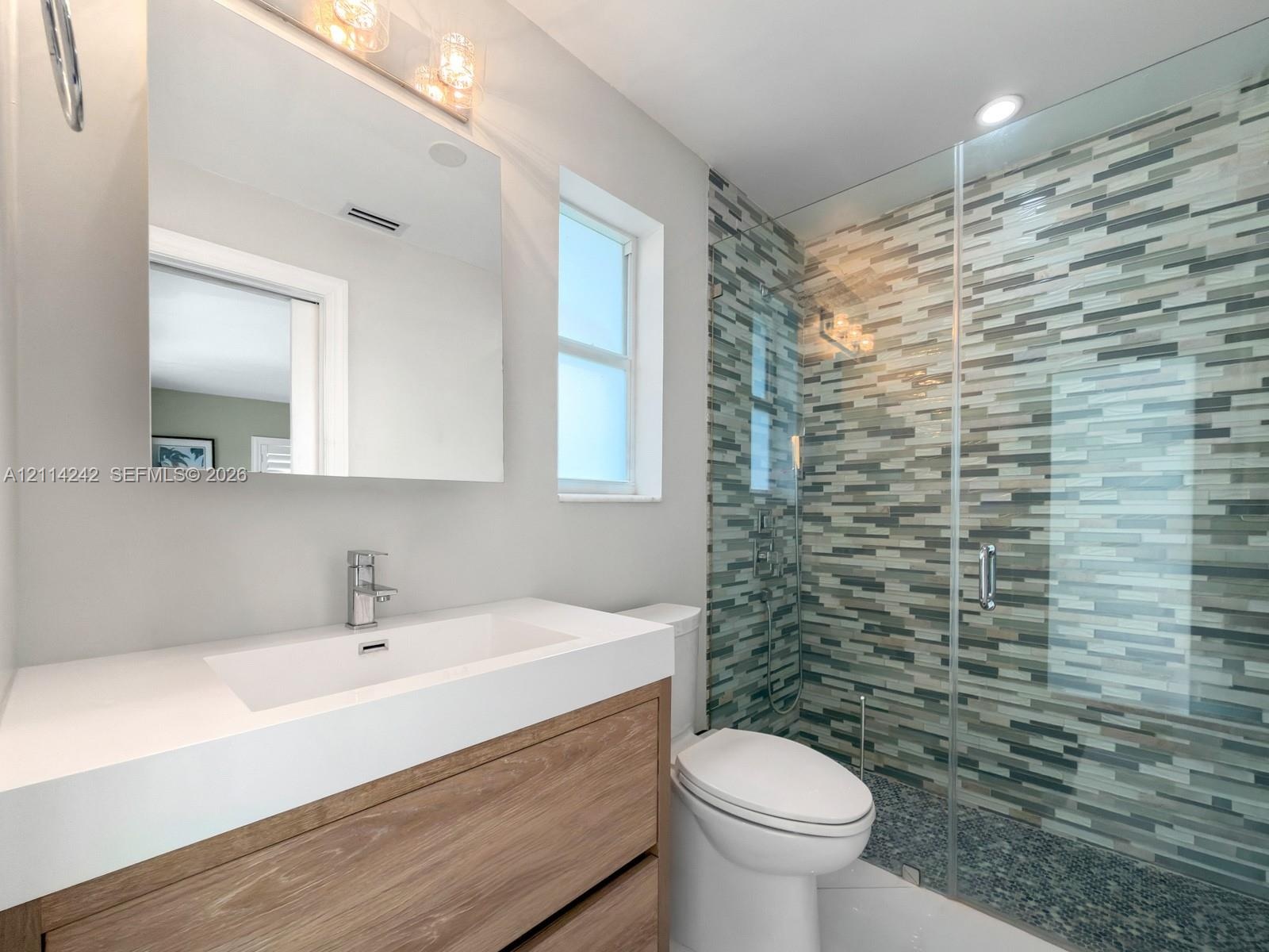 Wood-finish floating vanity with a white countertop and rectangular undermount sink, complemented by a mosaic tile walk-in shower featuring a glass en