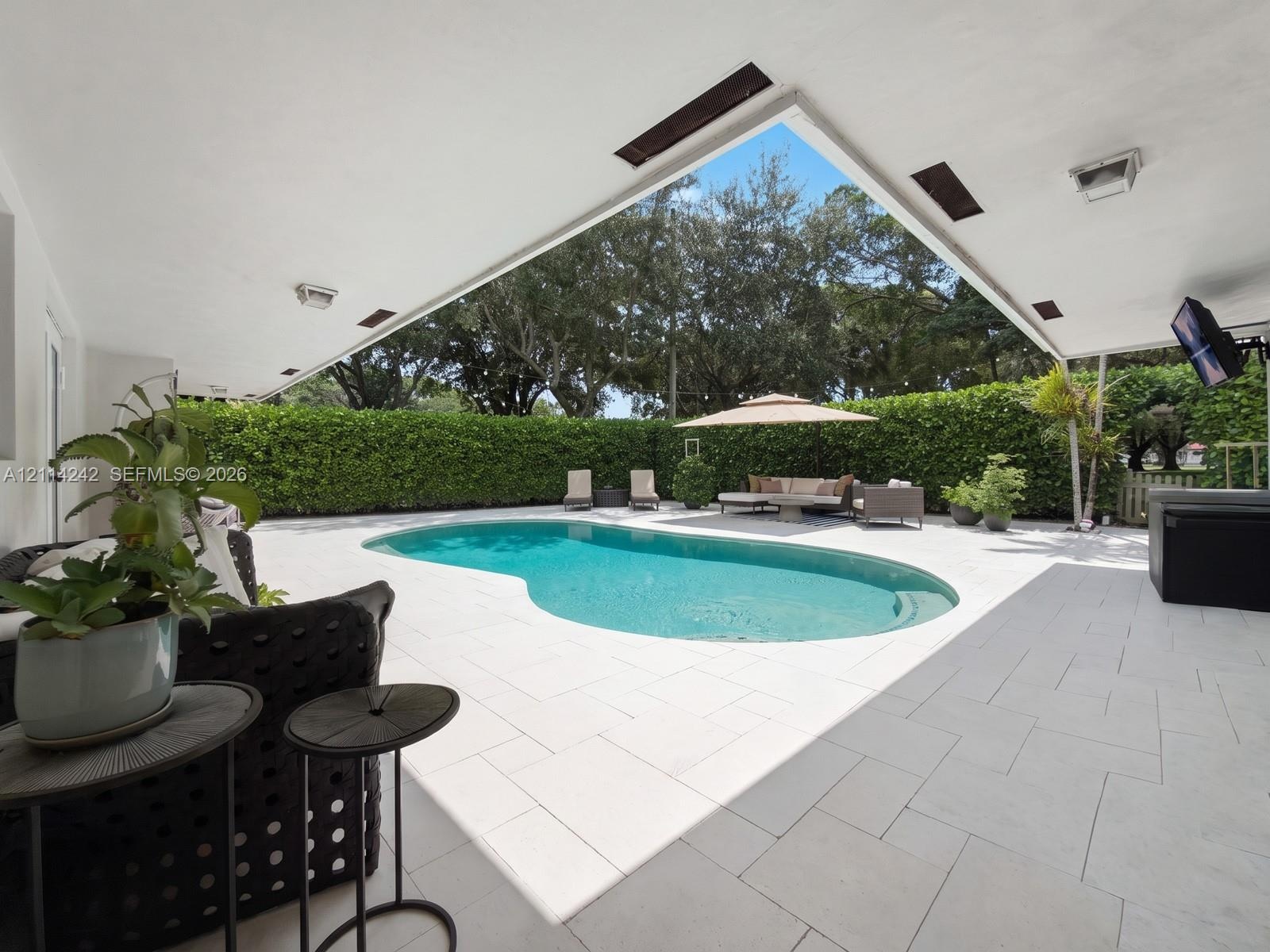 Covered outdoor patio with stone-finish tile flooring, recessed lighting, and a freeform swimming pool surrounded by a tall, mature privacy hedge