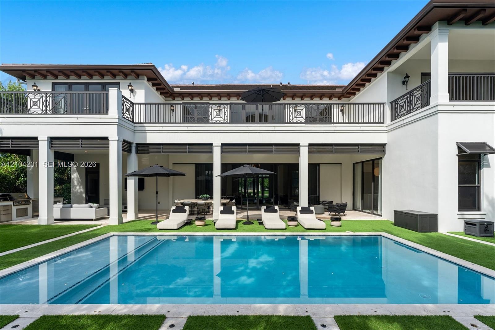Custom swimming pool with stone coping, covered patio, second-story balcony with metal railing, and built-in outdoor kitchen